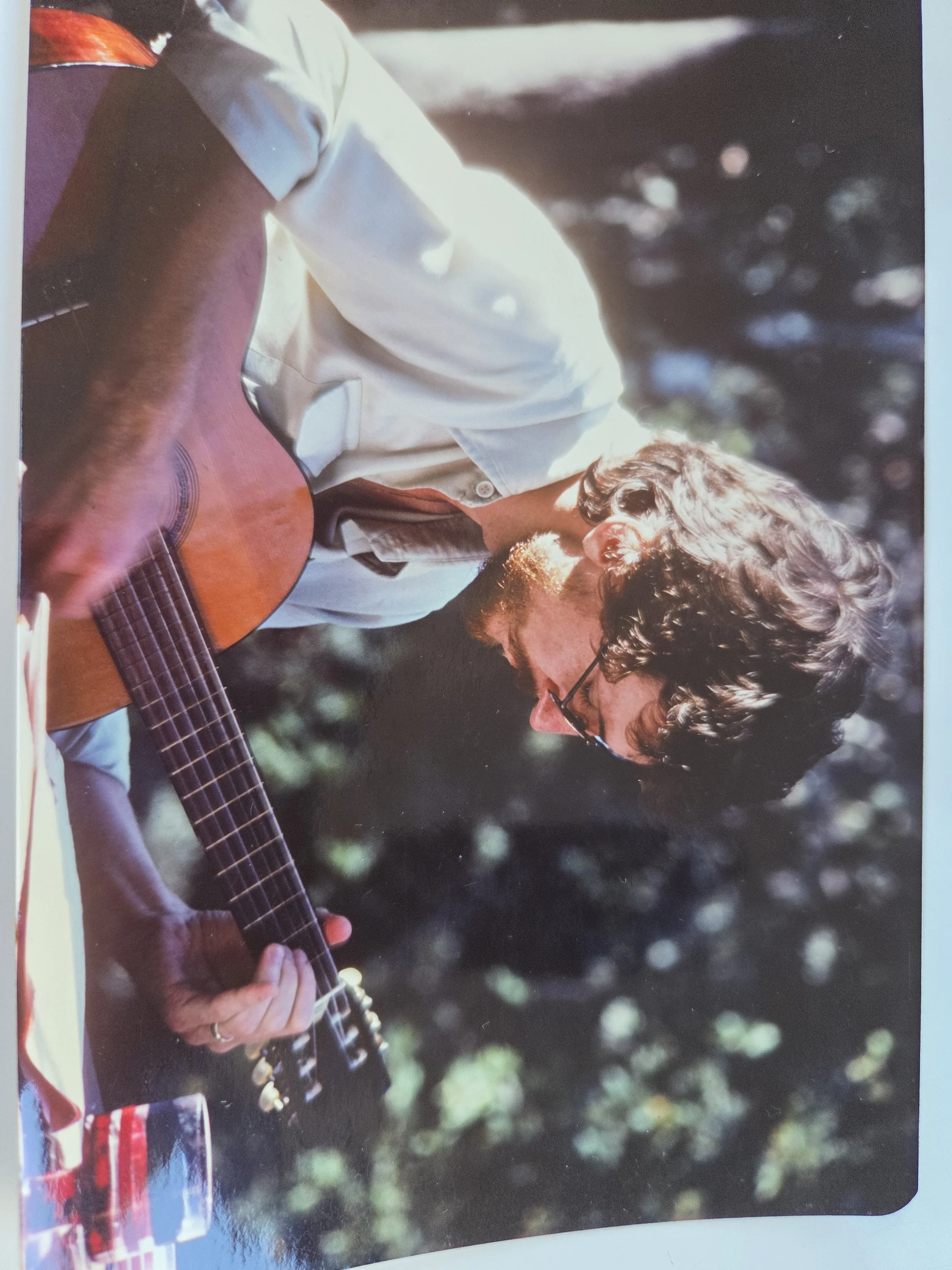 A man with curly hair and glasses playing an acoustic guitar outdoors with sunlight filtering through trees in the background.