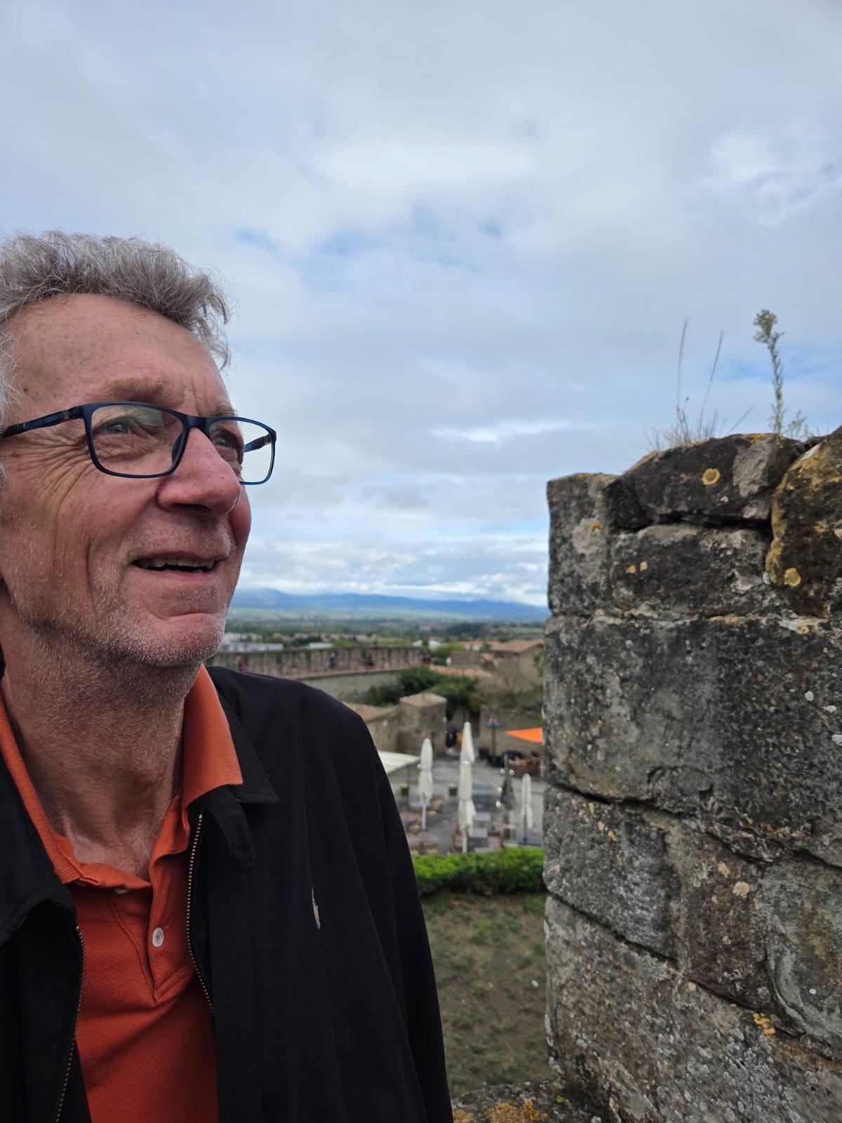 A smiling older man with glasses standing beside a stone wall, with a rural landscape and cloudy sky in the background.