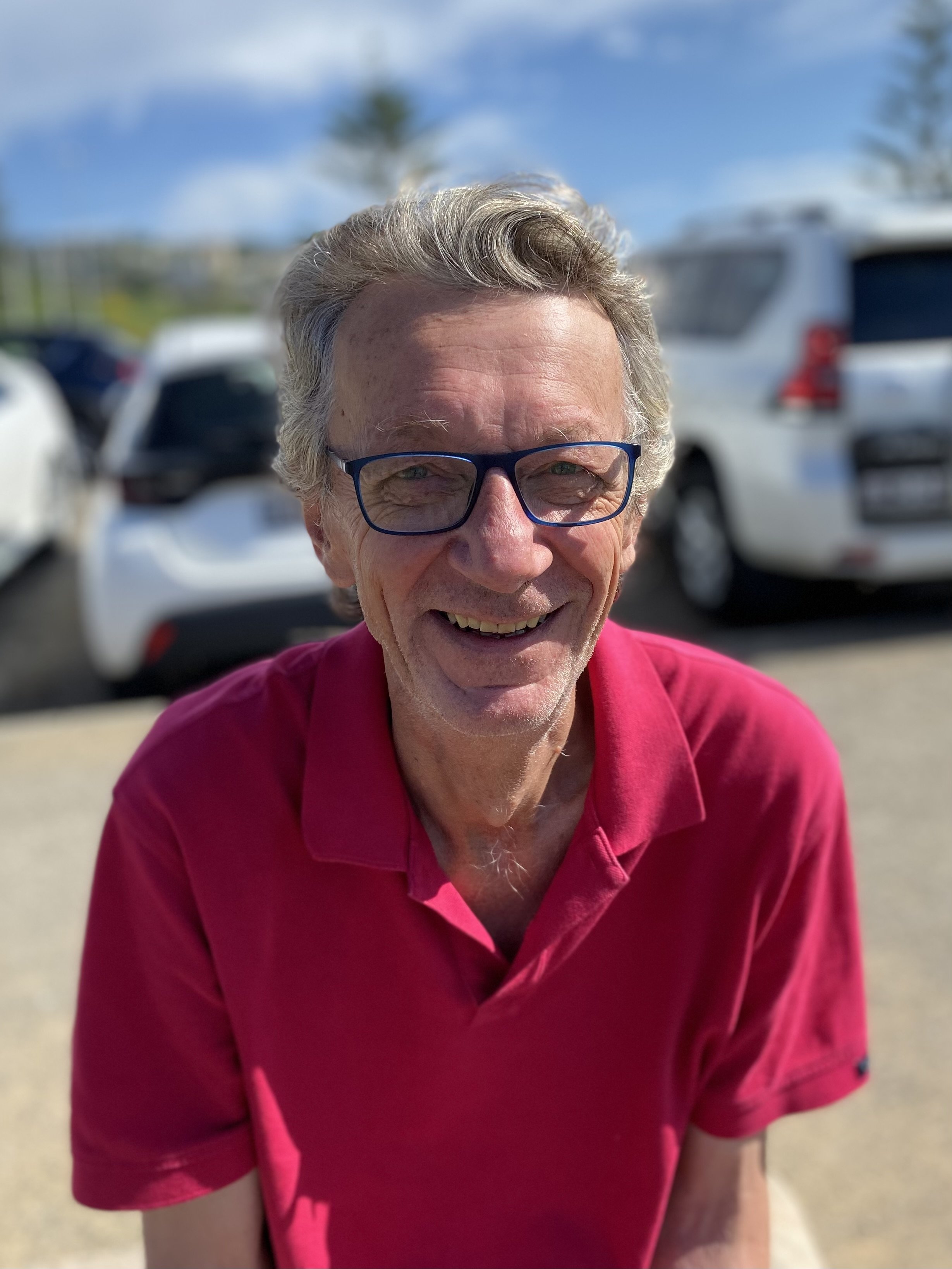 Close-up of a smiling elderly man wearing glasses and a pink polo shirt outdoors, with cars and trees in the blurred background.