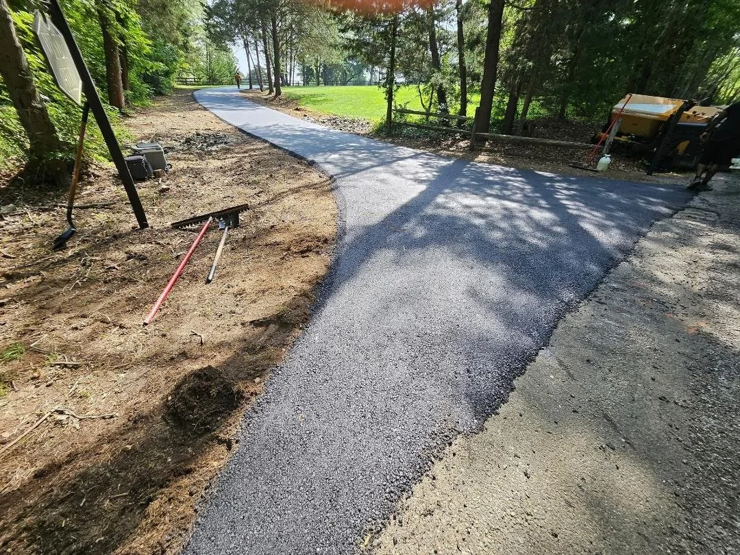 A paved sidewalk under construction with fresh asphalt, surrounded by trees and a grassy area. Construction tools and equipment are nearby.