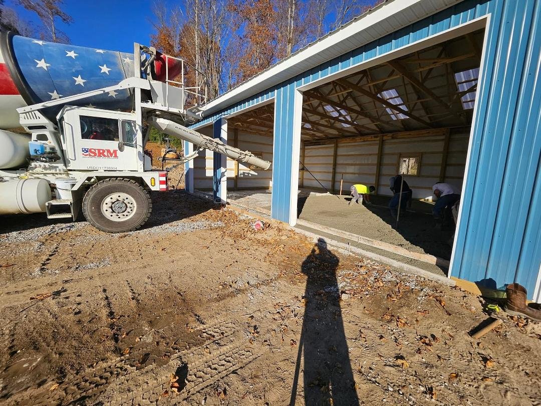 Construction workers are pouring concrete inside a blue metal building, with a cement mixer truck nearby preparing the foundation. The area is outdoors with a clear sky and some trees in the background.