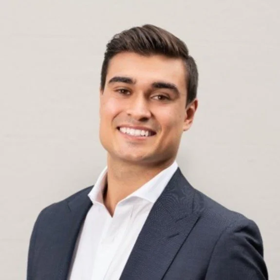 Smiling young man in a suit and white shirt against a plain light-colored background.