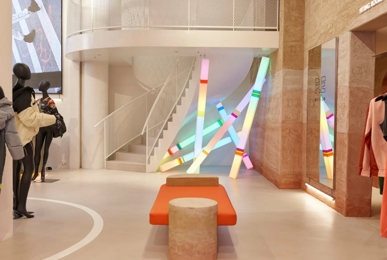 Interior of a retail store with mannequins dressed in athletic wear on the left, colorful neon light sculptures near a staircase in the center, a mirror on the right wall, and clothing racks.