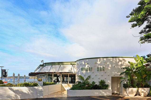 An outdoor bar with a white brick wall and large potted plants, with a waterfront view of a marina in the background and partly cloudy sky.