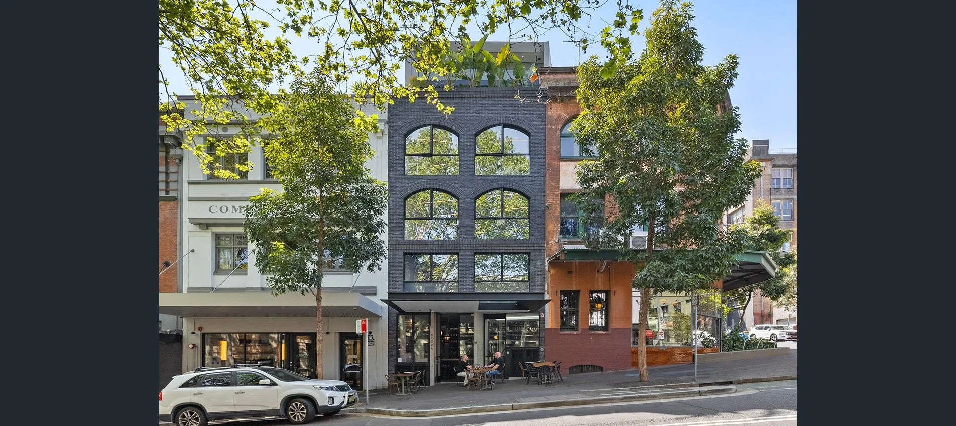 A city street scene with a modern three-story building featuring large arched windows, two trees lining the sidewalk, and parked cars. People sit at outdoor tables in front of the building.