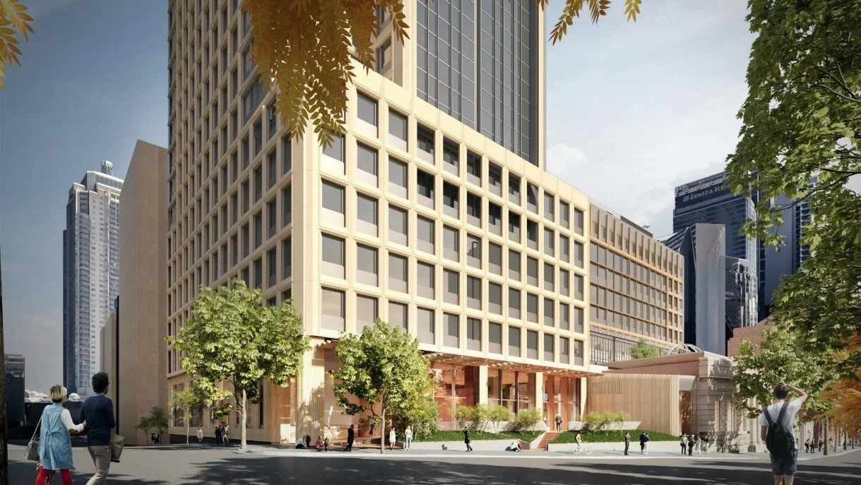 A modern cityscape showing a tall office building with a grid-like facade, trees lining the street, and people walking on the sidewalk.