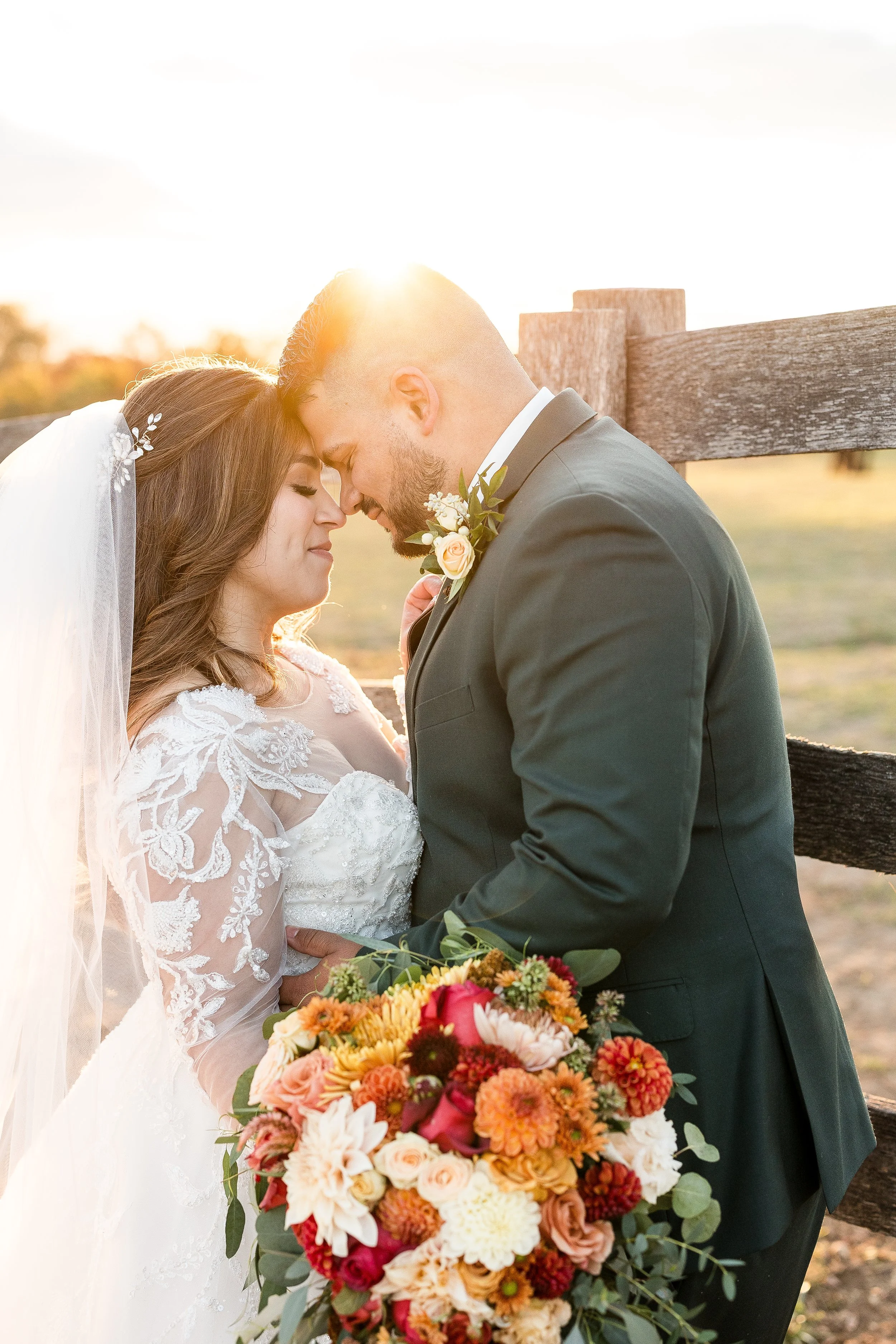 Bride and groom touching foreheads beside a wooden fence during golden-hour wedding portraits
