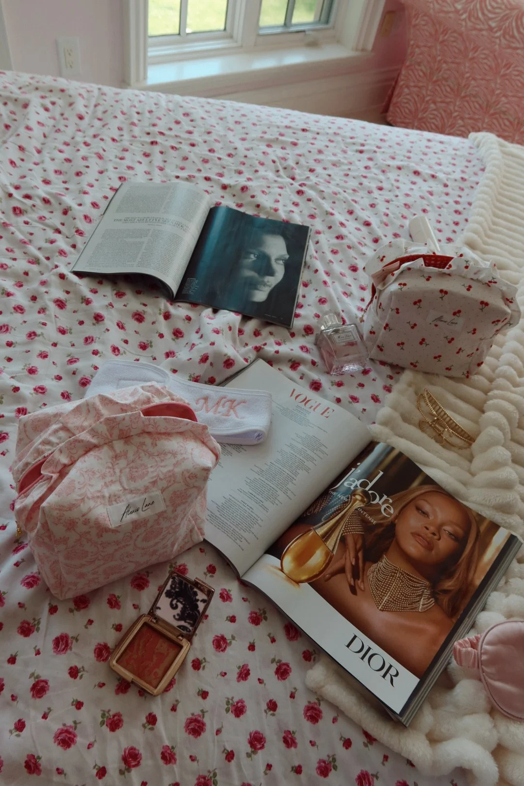 A bed with a pink floral bedspread, with a magazine, a book, a compact, a tissue box, and toiletries scattered on it. There is a window in the background.