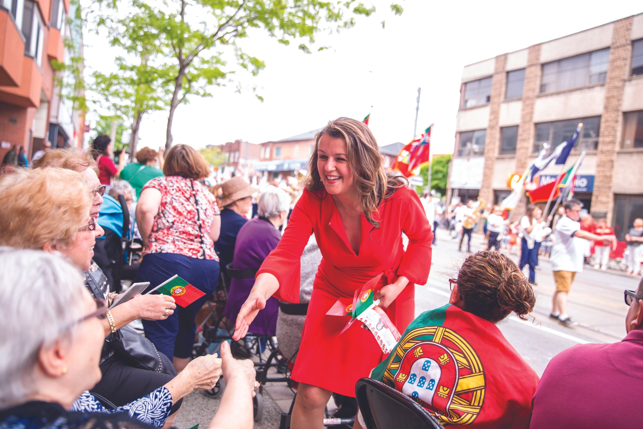Julie Dzerowicz in a red dress smiling and shaking hands with an elderly woman at an outdoor event, with other elderly people and portuguese flags in the background.