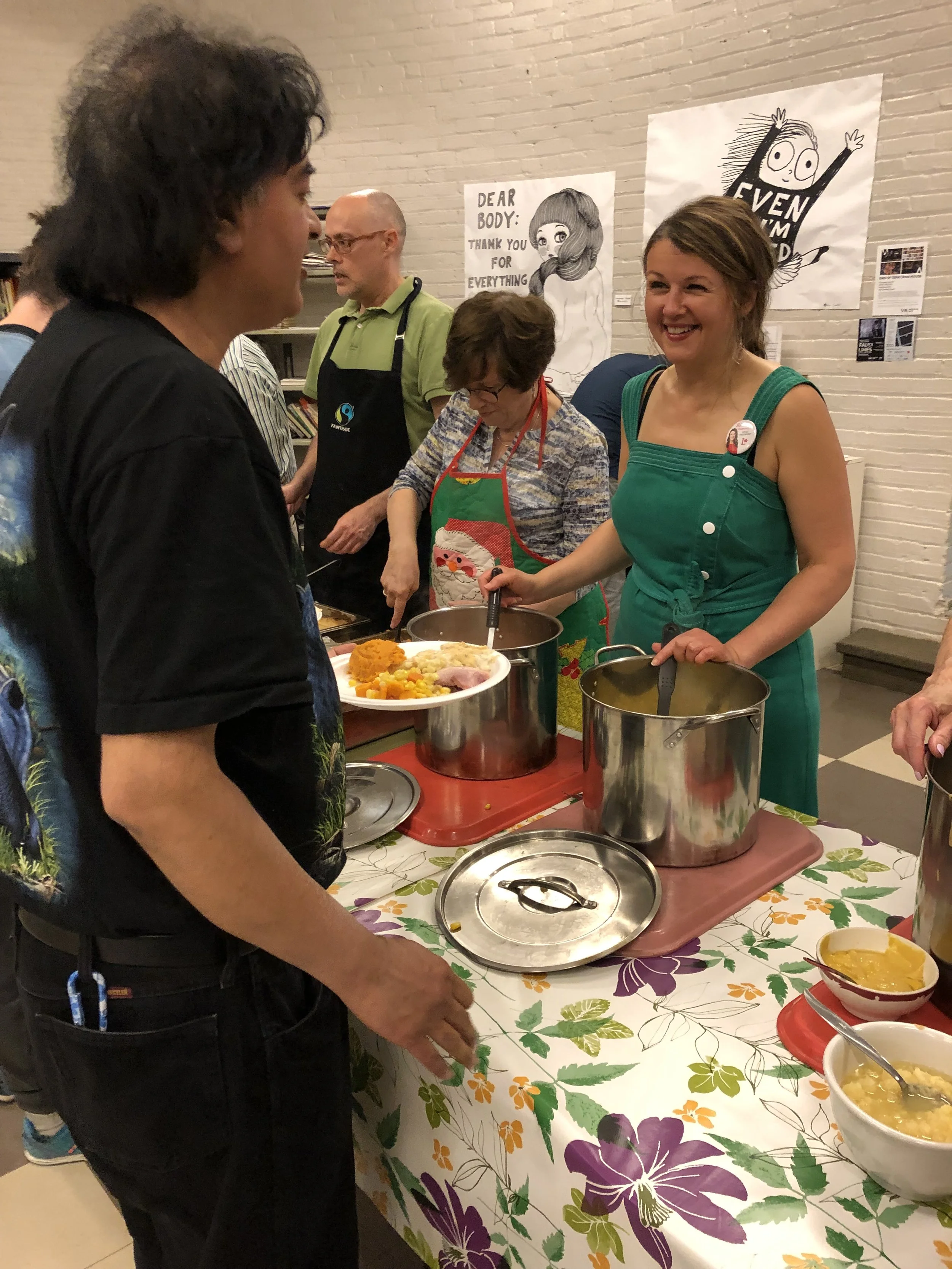 Julie Dzerowicz and other volunteers serving food at a holiday-themed gathering with bowls and a plate of food on a table covered with a floral tablecloth.