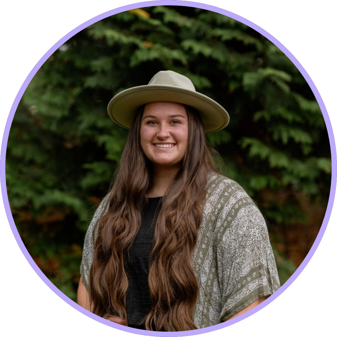 Emily Burns standing in front of pine trees, smiling at the camera with her long brown hair, a green shawl and a green western-style hat.