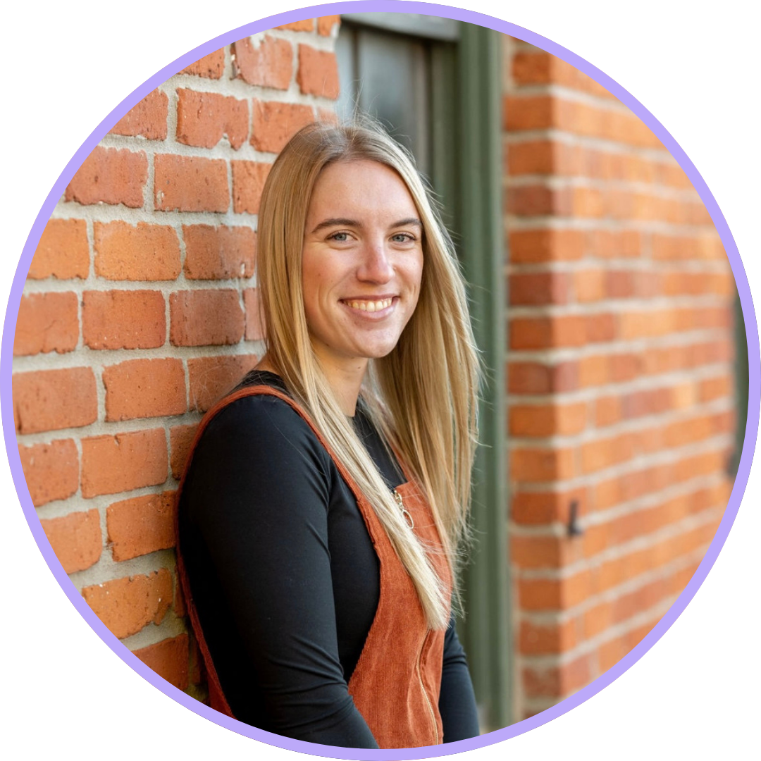 Hailey Ratliff, leaning against a brick wall with long, straight blonde hair, wearing a black long sleeve, a teracotta-colored dress and a bright smile.