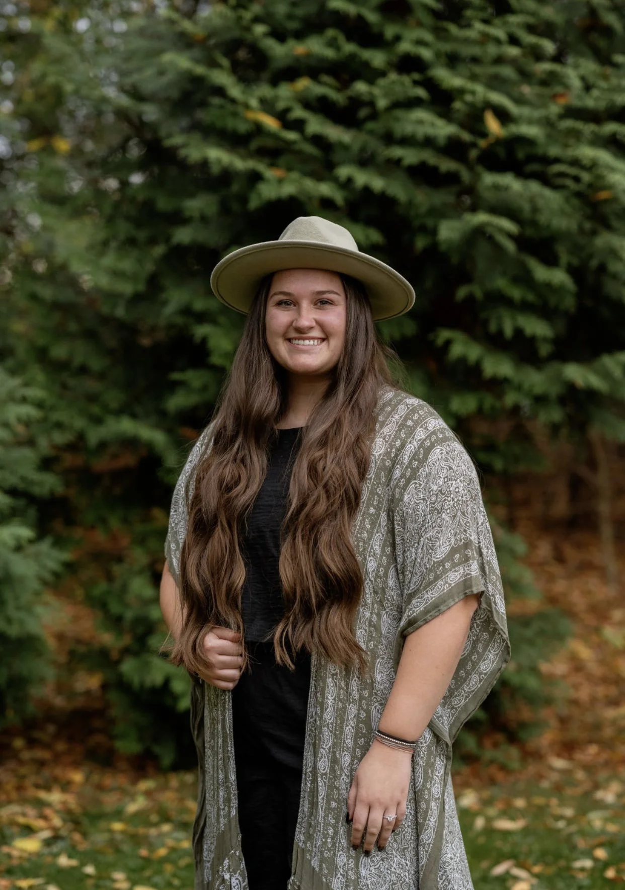 Emily Burns standing in front of pine trees, smiling at the camera with her long brown hair, a green shawl and a green western-style hat.