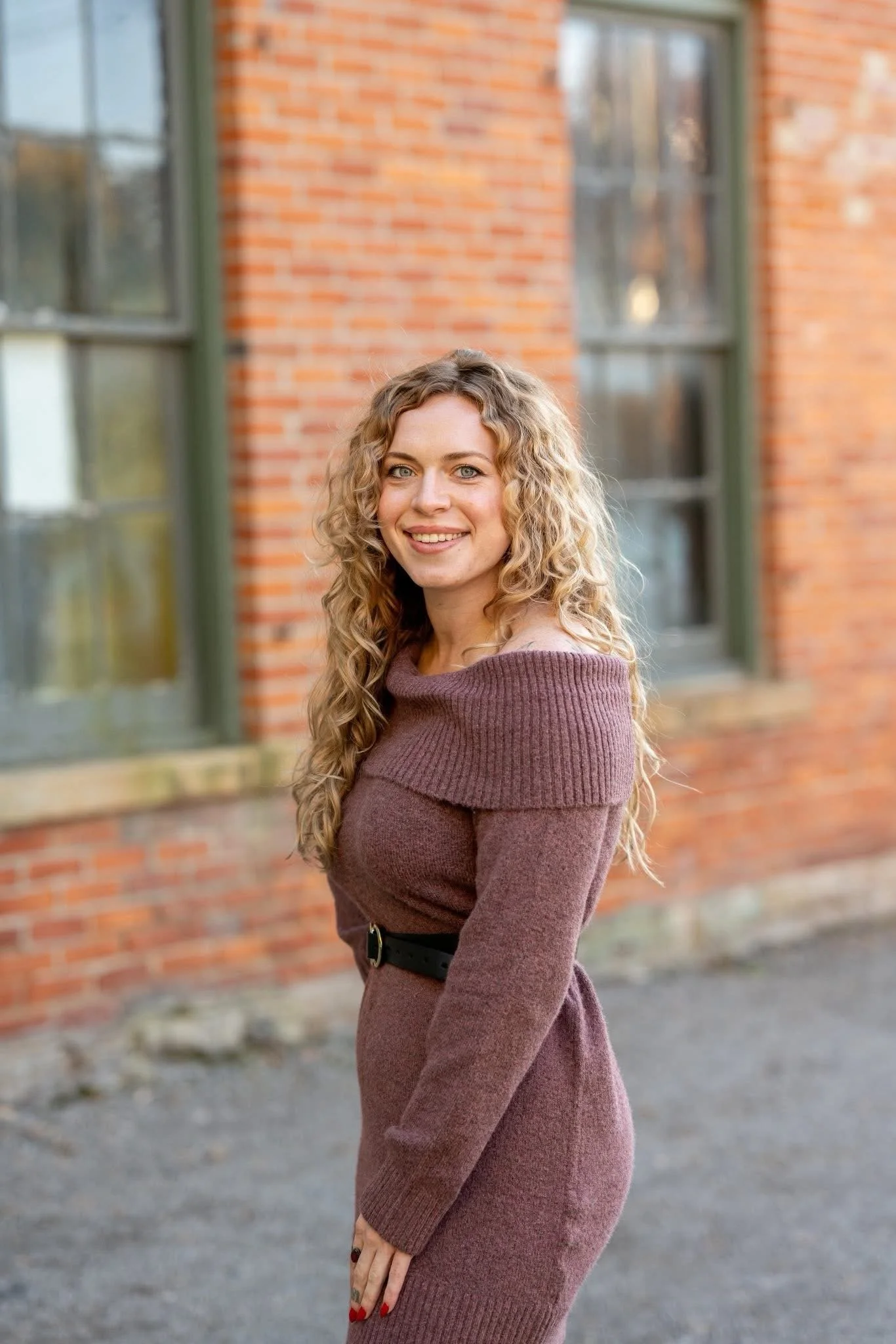Hannah McMillen standing in front of a brick building, smiling at the camera with curly hair and a burgundy colored off-the-shoulder dress.