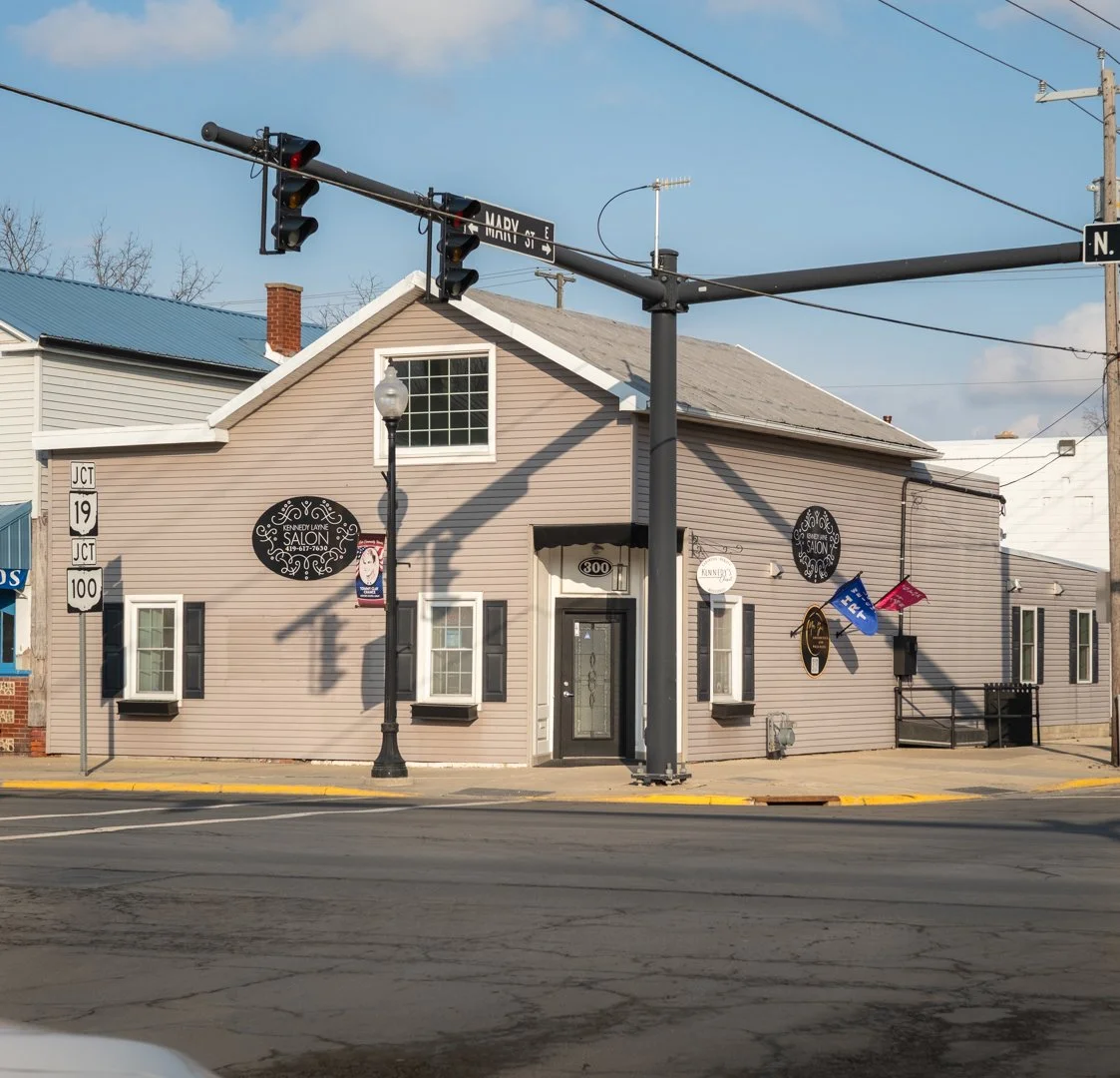 A two-story building with beige siding and black shutters, located at the corner of an intersection. The building has a sign that reads 'Kennedy Lane Salon.' There are street signs, a traffic light, and power lines visible. The sky is clear with some clouds.
