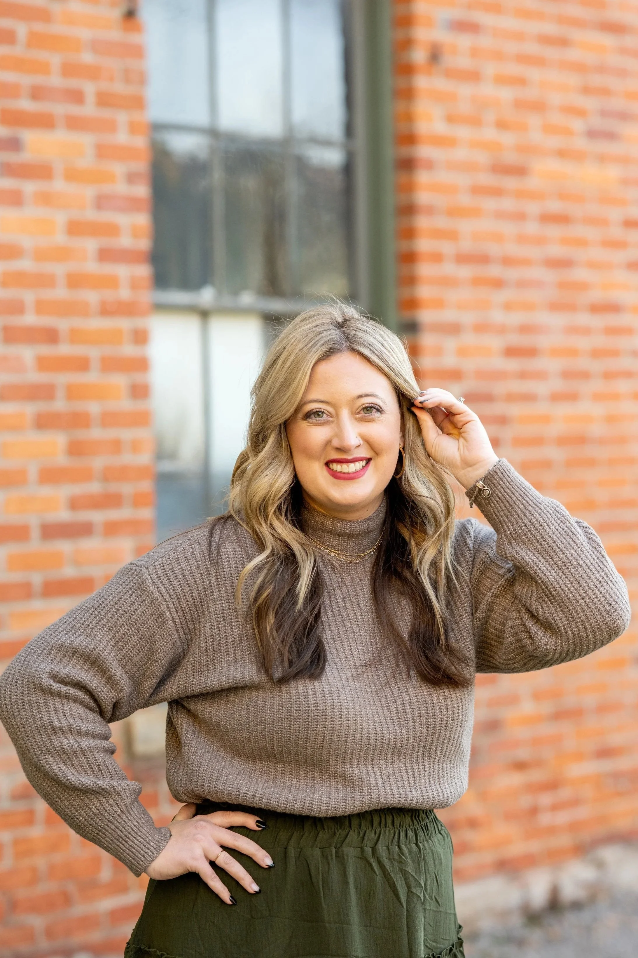 Erin Hair, wearing a brown sweater with blonde and brown curled hair and a bright smile.