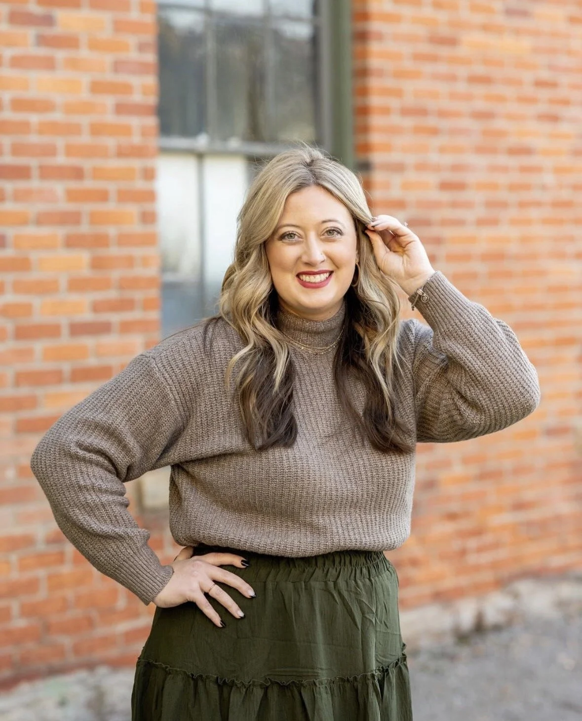 Erin Hair standing in front of a brick building, smiling at the camera with blonde and brown hair, a taupe sweater and green skirt.