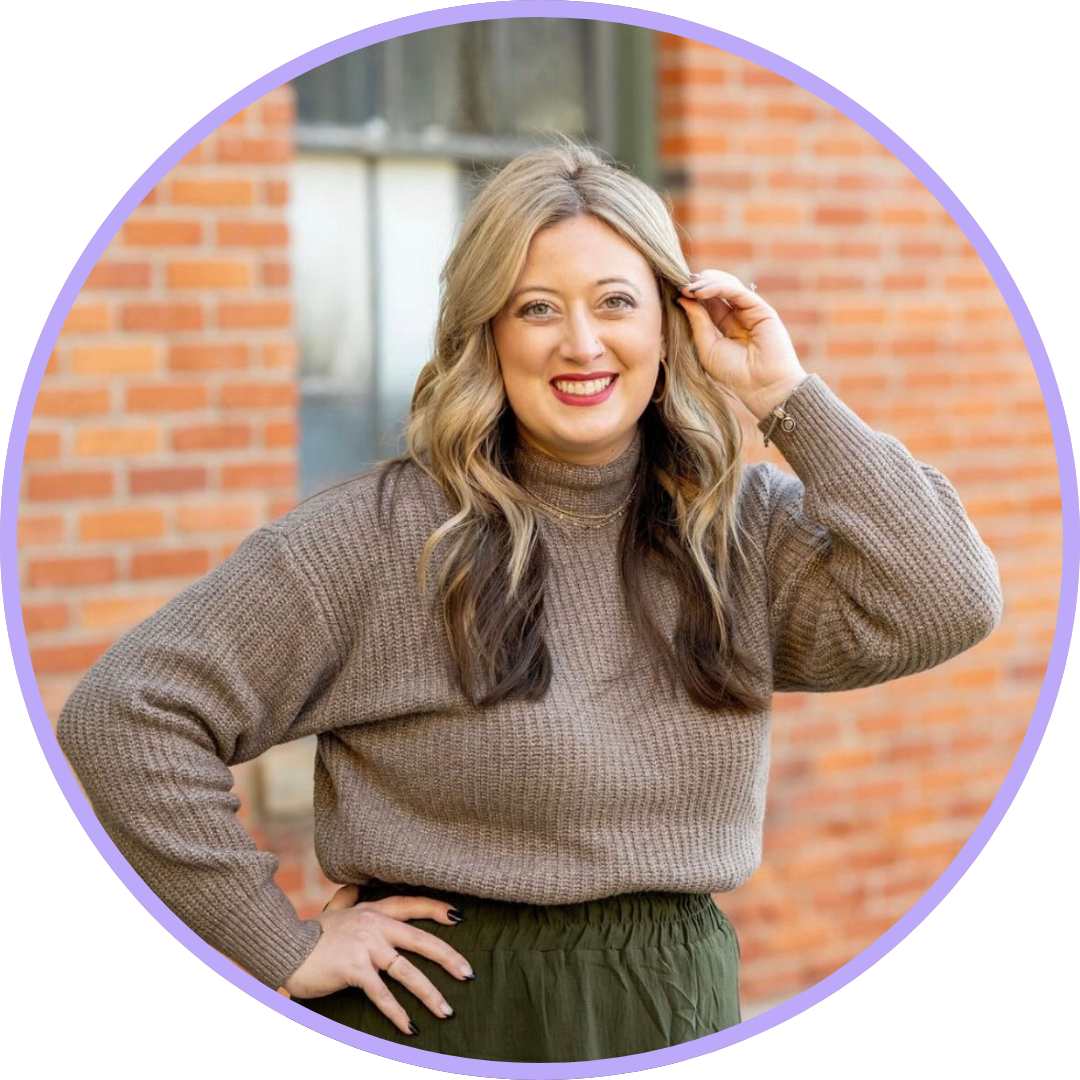 Erin Hair standing in front of a brick building, smiling at the camera with blonde and brown hair, a taupe sweater and green skirt.