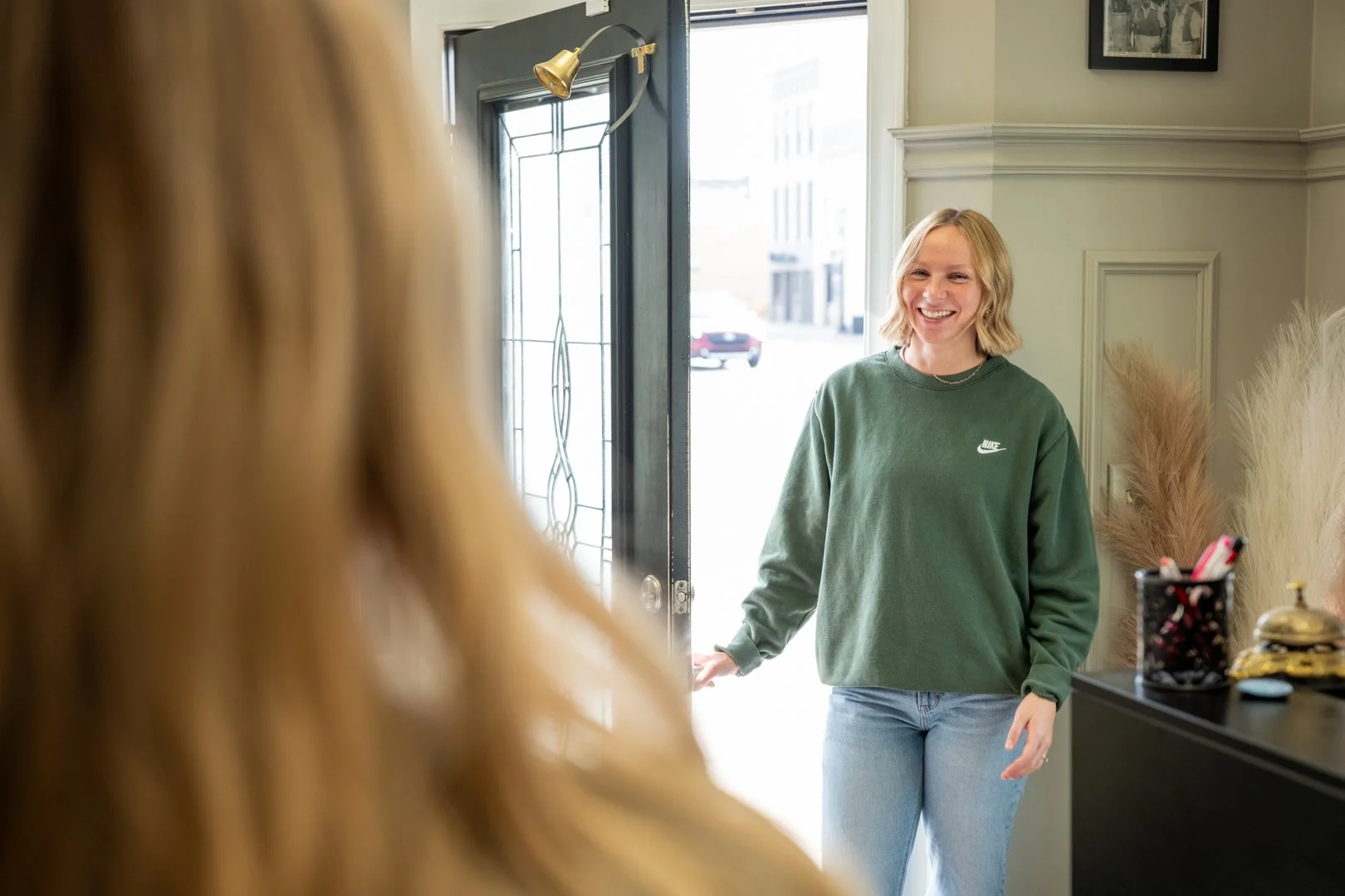 A woman with blonde hair and a green Nike sweatshirt smiling at another person with long blonde hair standing inside Kennedy Layne Salon near the entrance.