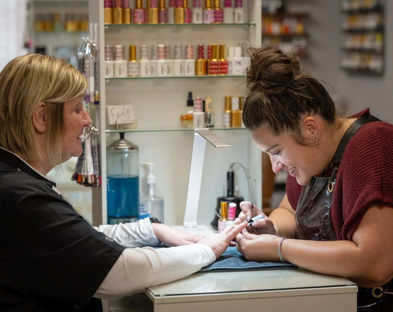 A nail technician gives a manicure to a customer in a salon, with shelves of nail polish bottles in the background.