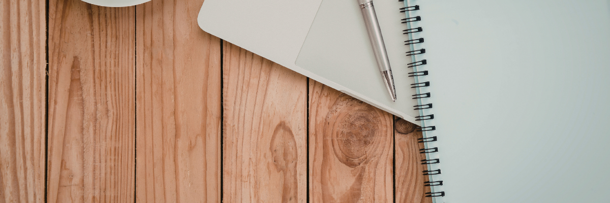 Top-down view of a wooden desk with a notebook, paper and pen.