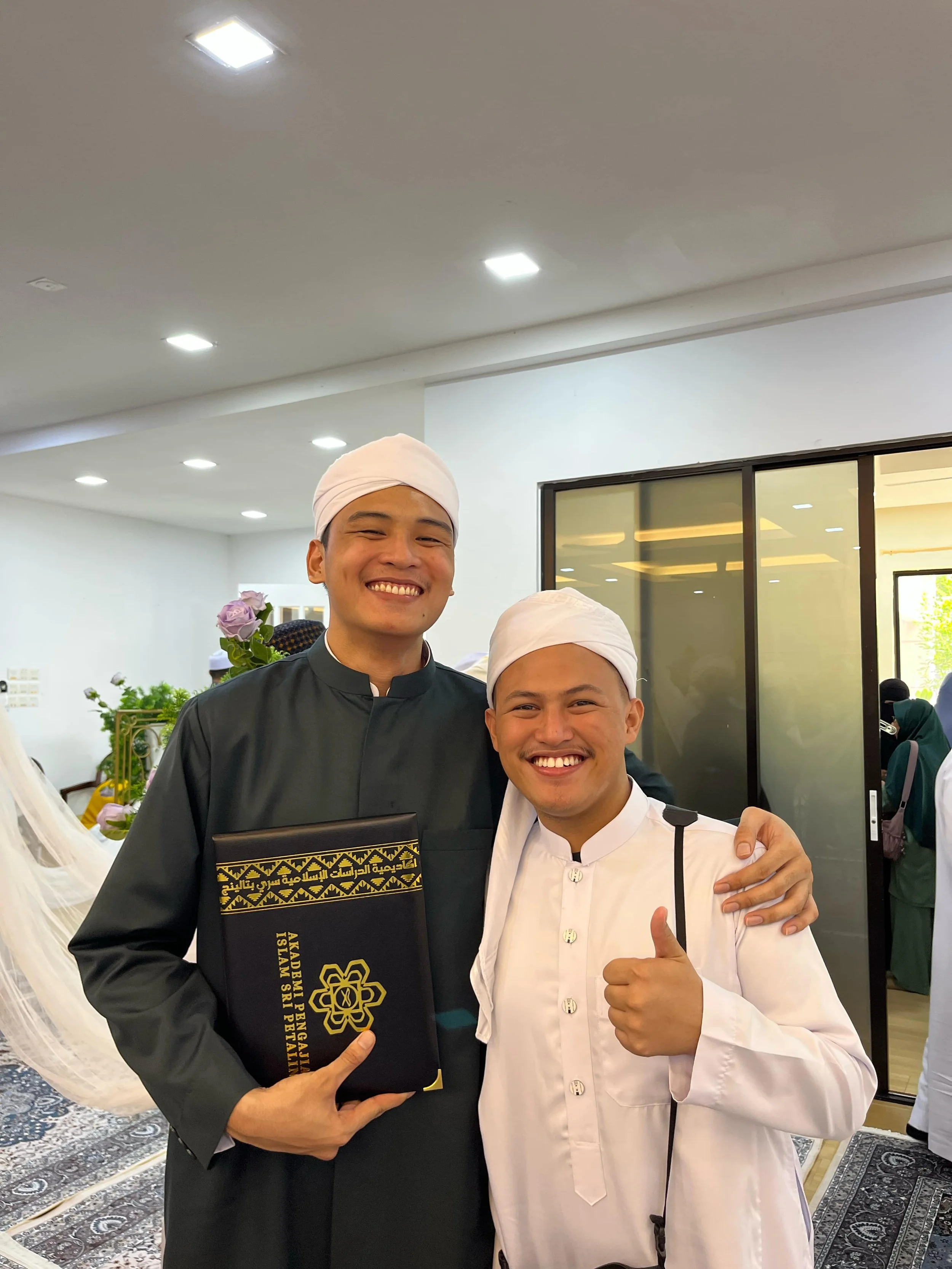 Two young men in traditional attire smiling and posing inside a decorated room, one holding a diploma or certificate, with floral arrangements and a glass window in the background.