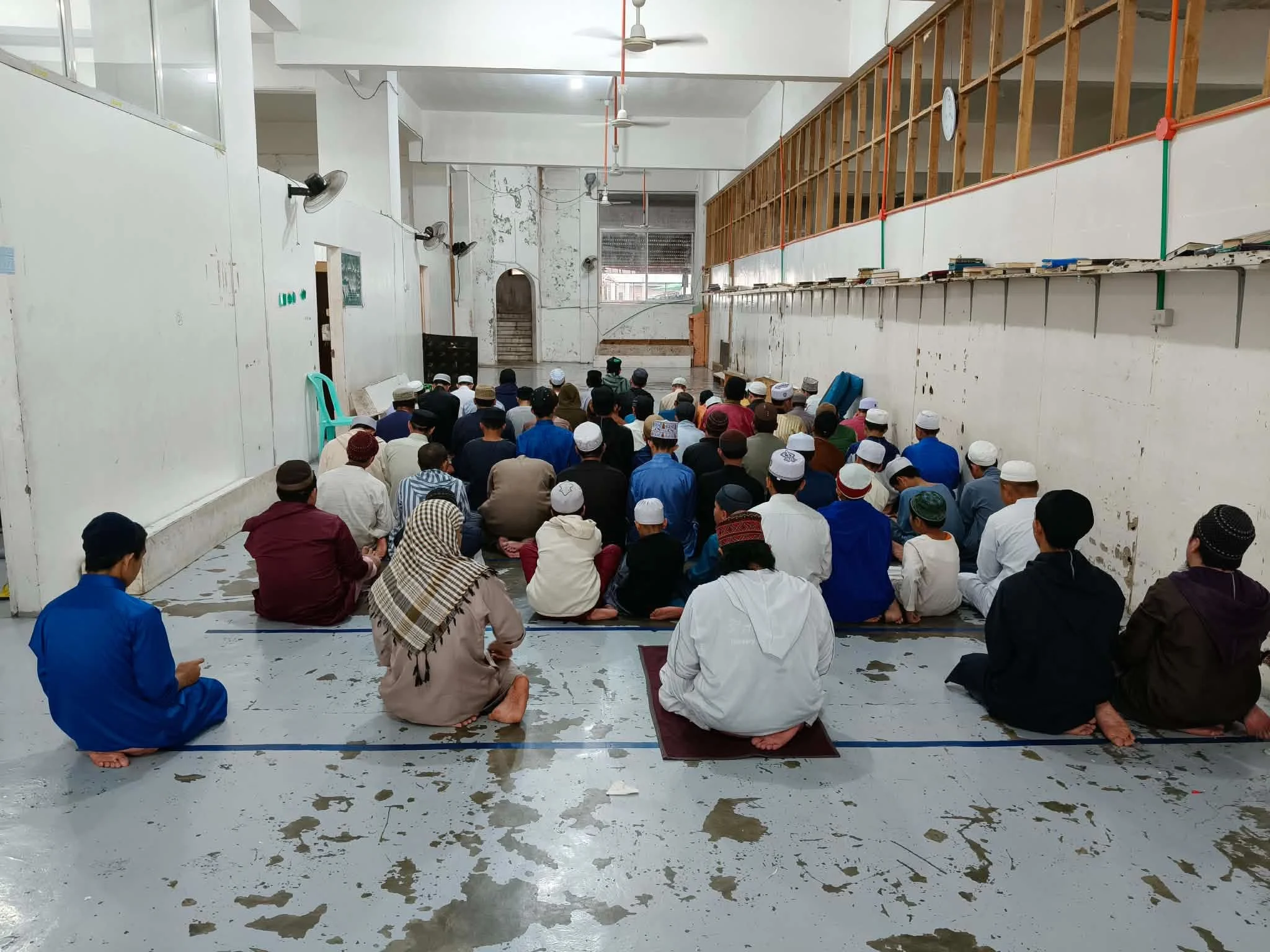 A large group of people, dressed in traditional clothing and white hats, are sitting on the floor facing forward in a spacious hall, possibly for prayer or a gathering.