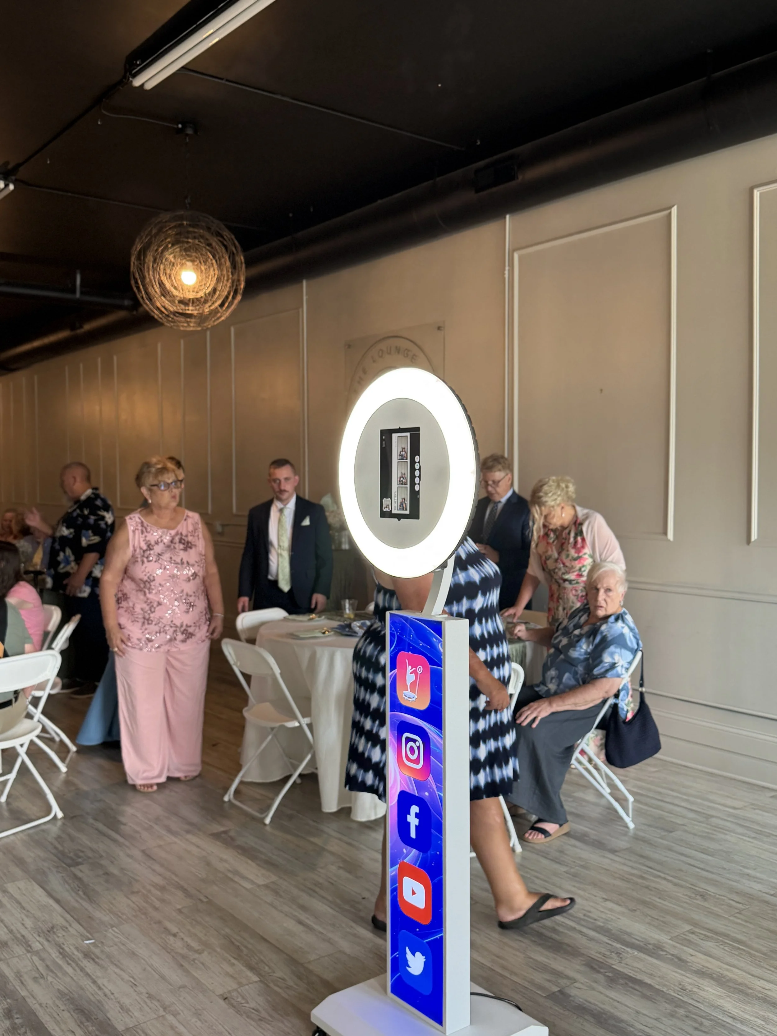 A social media photo booth with a circular light frame displaying app icons including Instagram, Facebook, YouTube, and Twitter at a social gathering, with elderly people and a woman in a pink dress in the background.