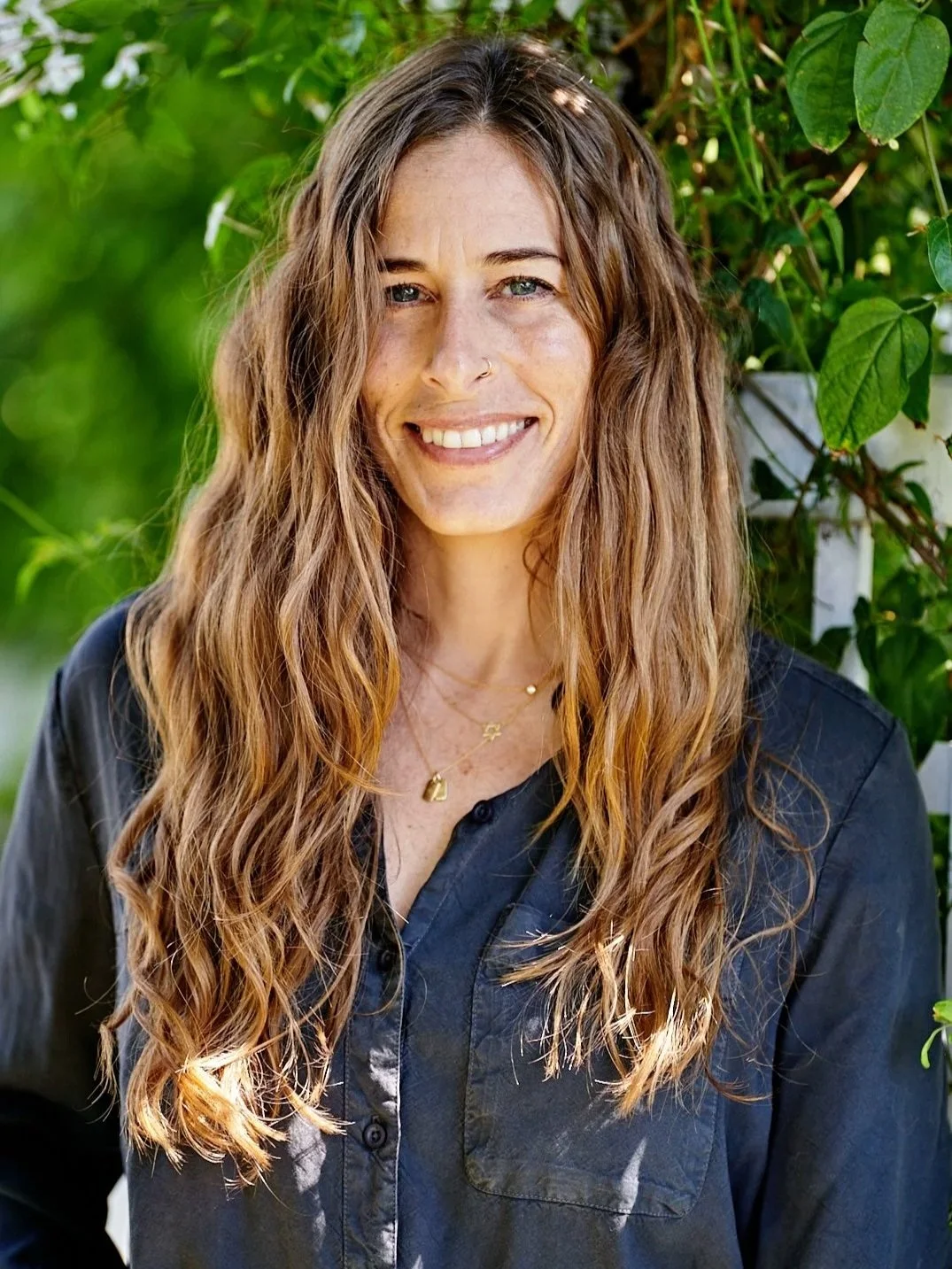 A woman with long, wavy, light brown hair and a nose piercing smiling outdoors with green foliage in the background, wearing a dark button-up shirt and layered necklaces.