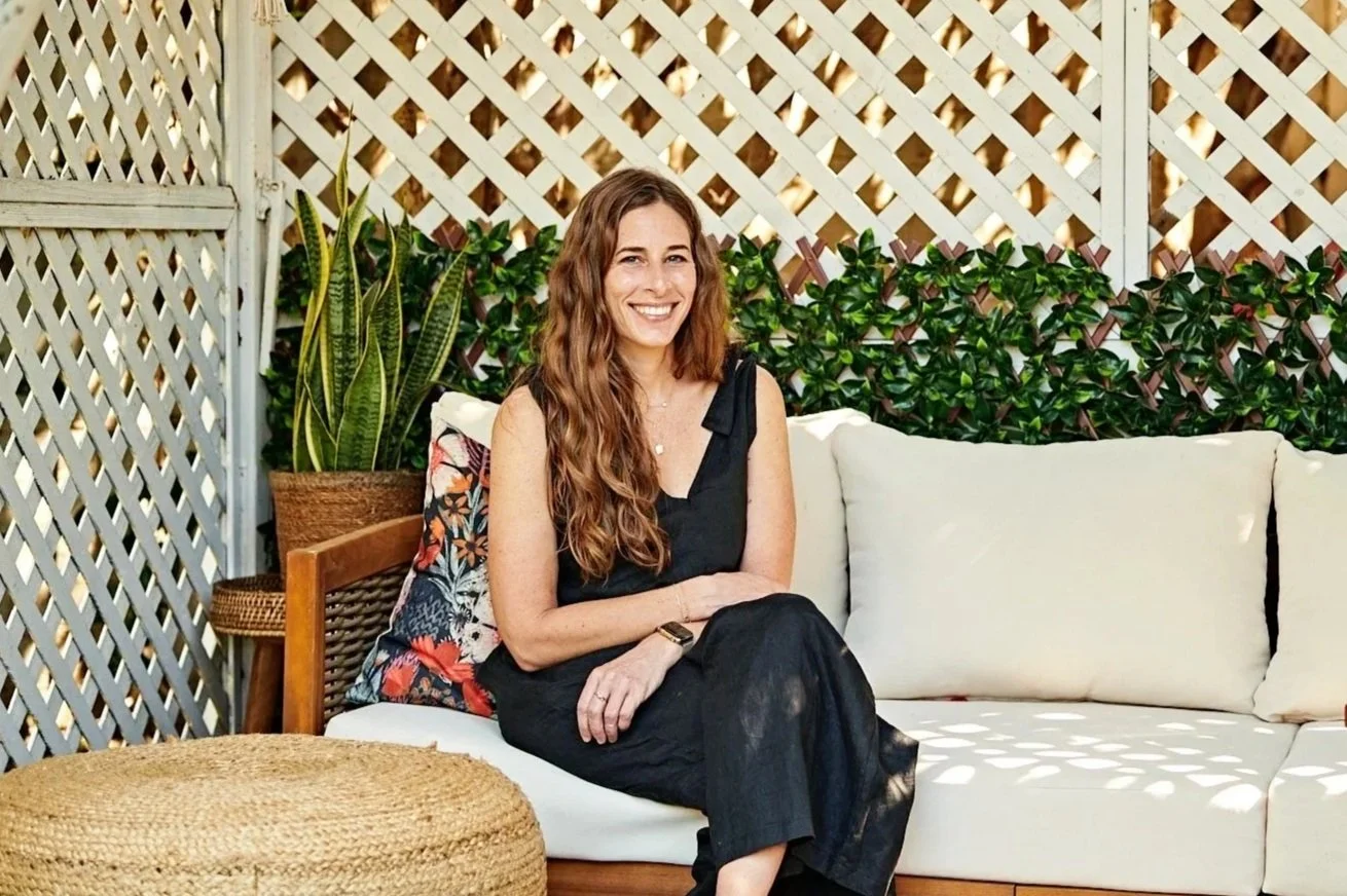A woman with long wavy hair sitting on a white outdoor sofa, smiling, surrounded by plants and a lattice fence in a backyard setting.
