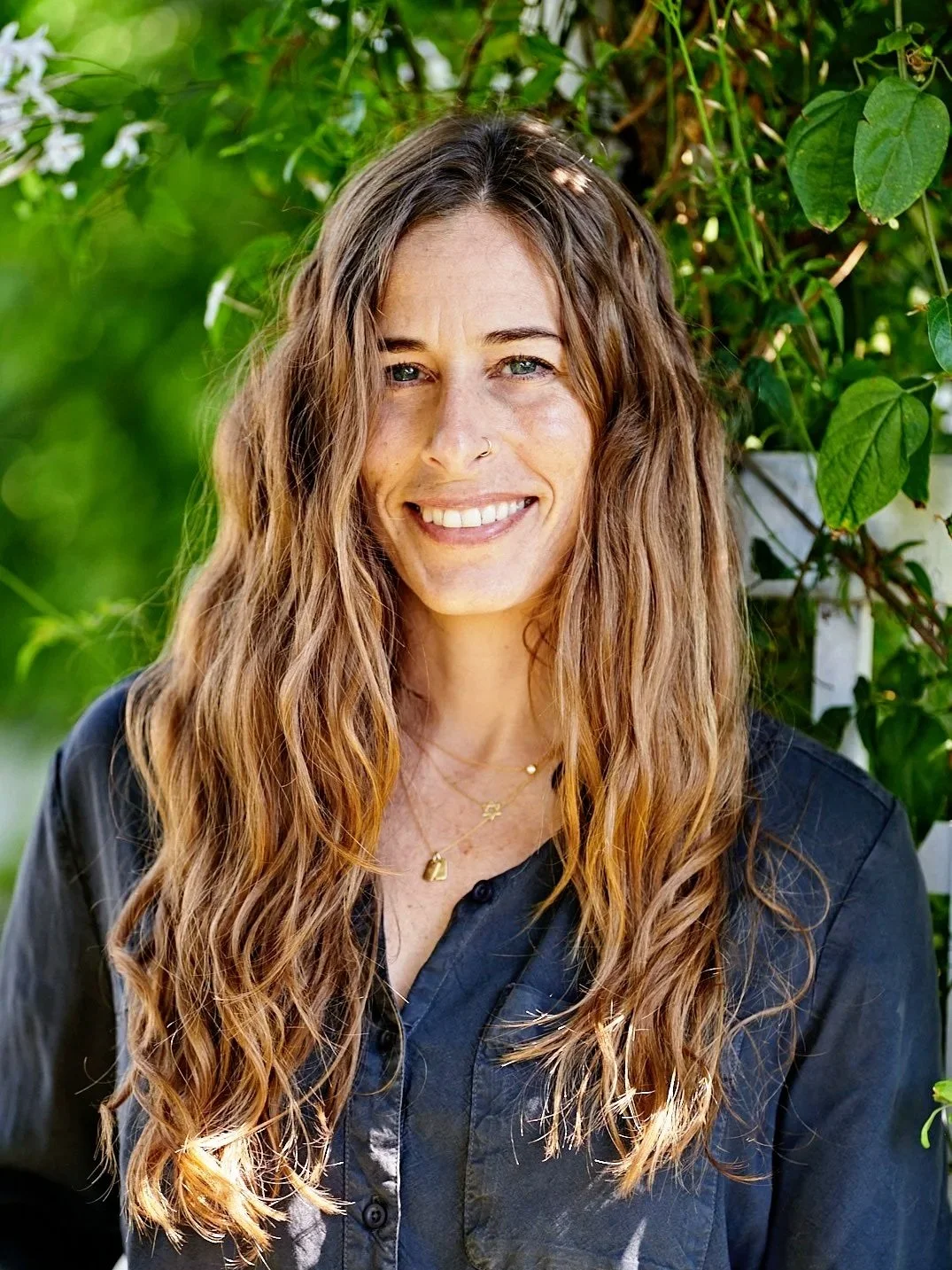 A woman with long, wavy brown hair smiling outdoors with green foliage in the background.