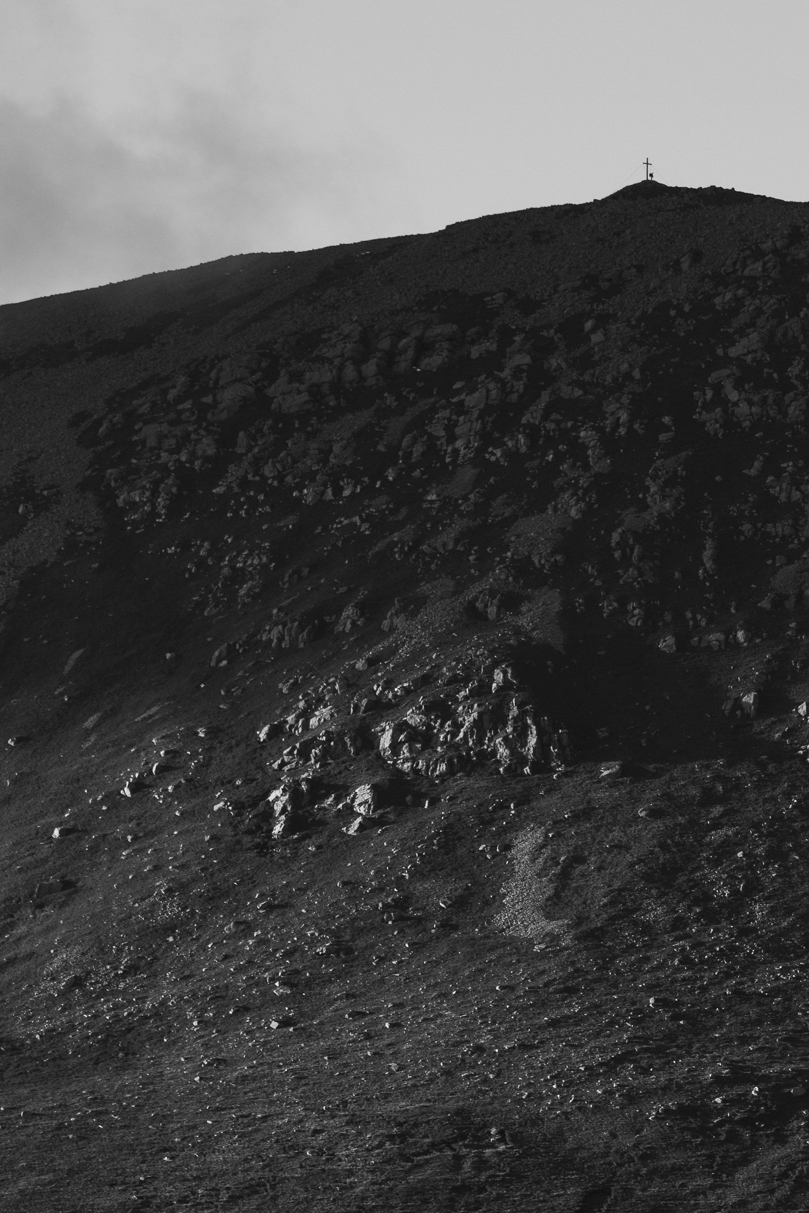 Black and white photo of a rocky mountain landscape with a cross and a small figure at the mountain peak.