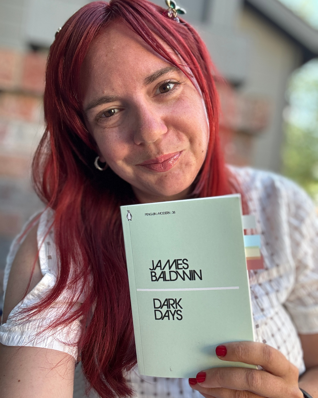 A woman with red hair holding a green book titled 'Dark Days' by James Baldwin, smiling at the camera.
