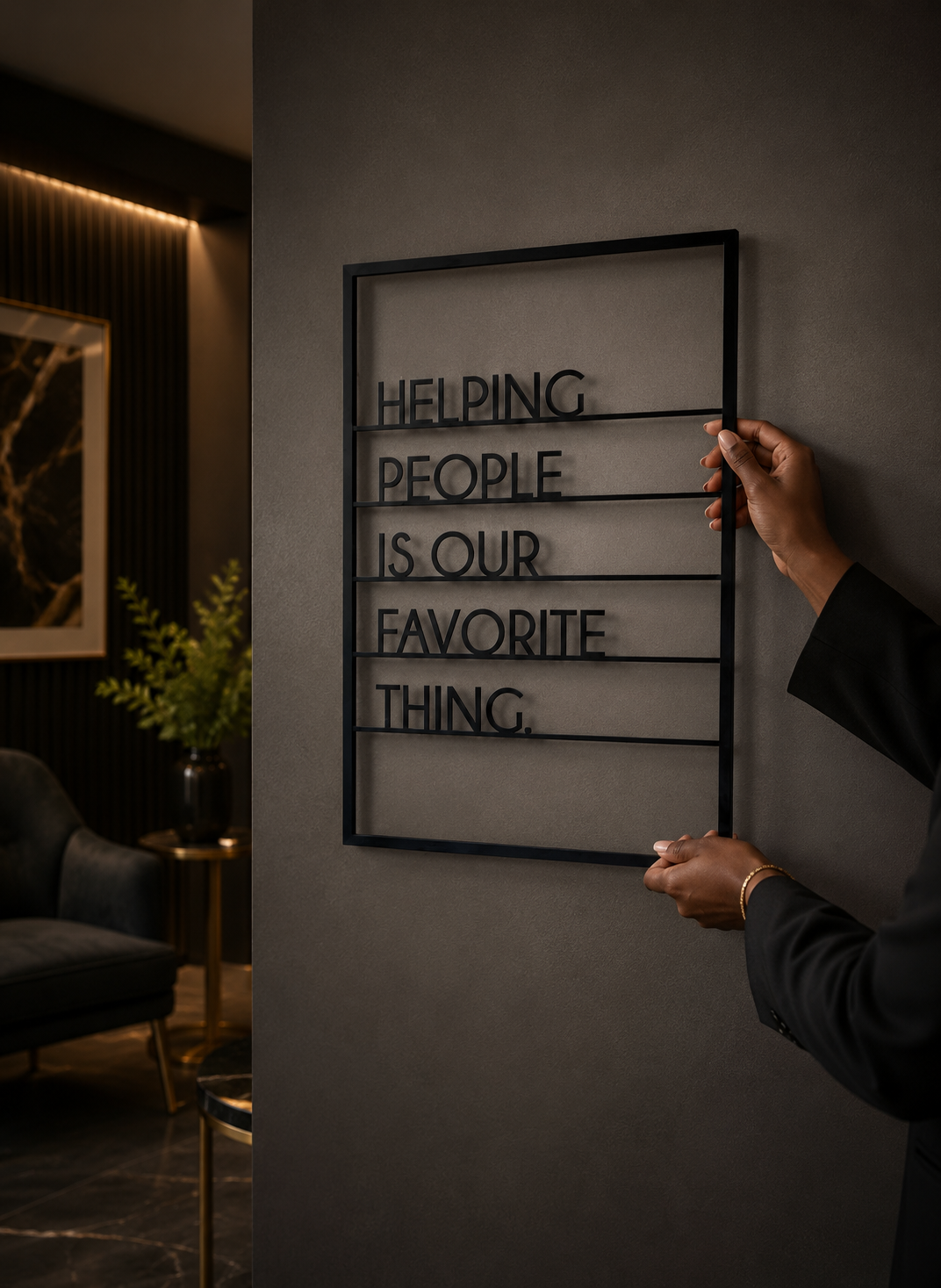 Person hanging a black metal framed sign on a gray wall that reads, "Helping people is our favorite thing." The sign has horizontal lines dividing the text into separate sections.