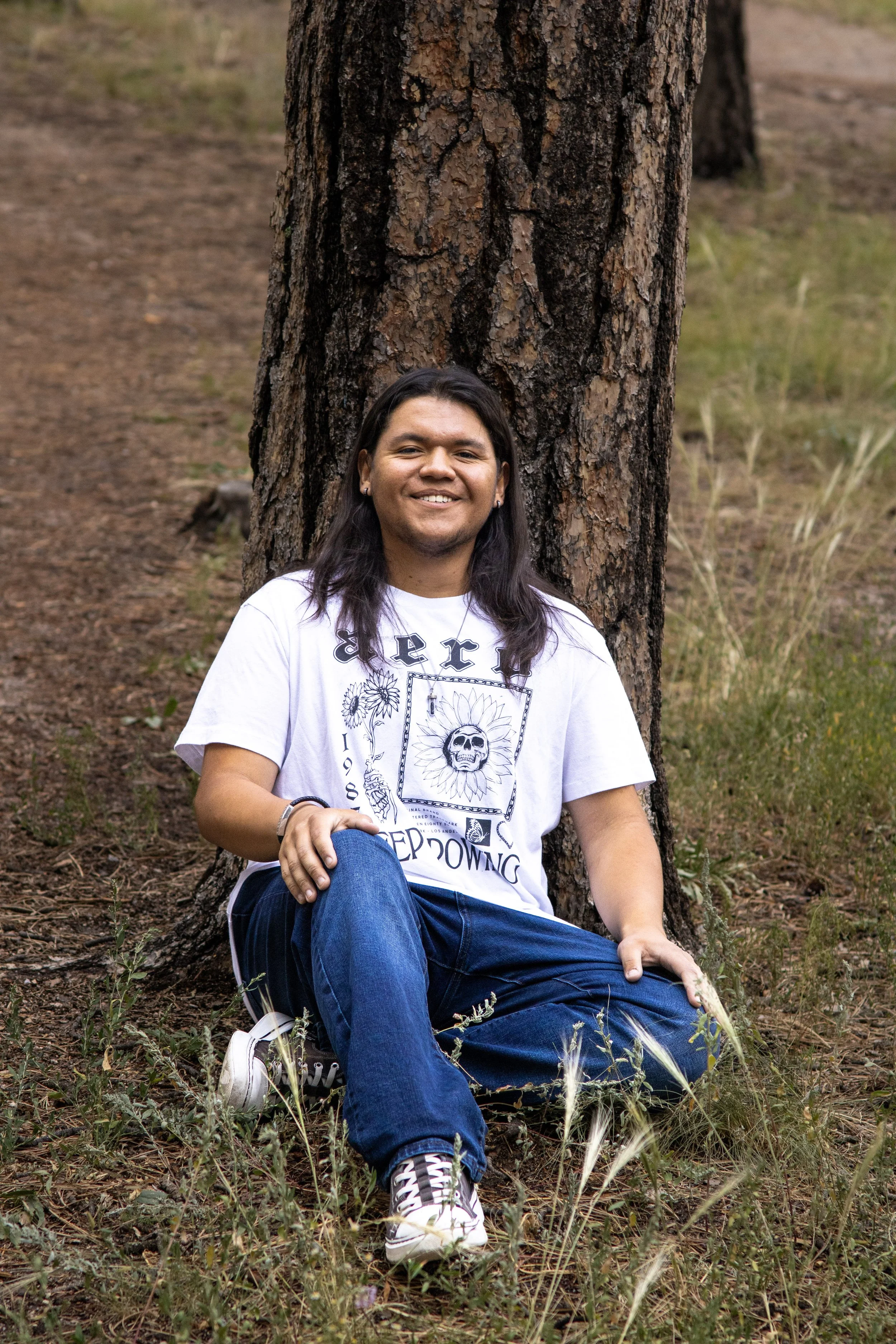A man with long dark hair, wearing a white graphic t-shirt, blue jeans, and sneakers, is sitting on the ground with his back against a tree in a natural outdoor setting with grass and soil.