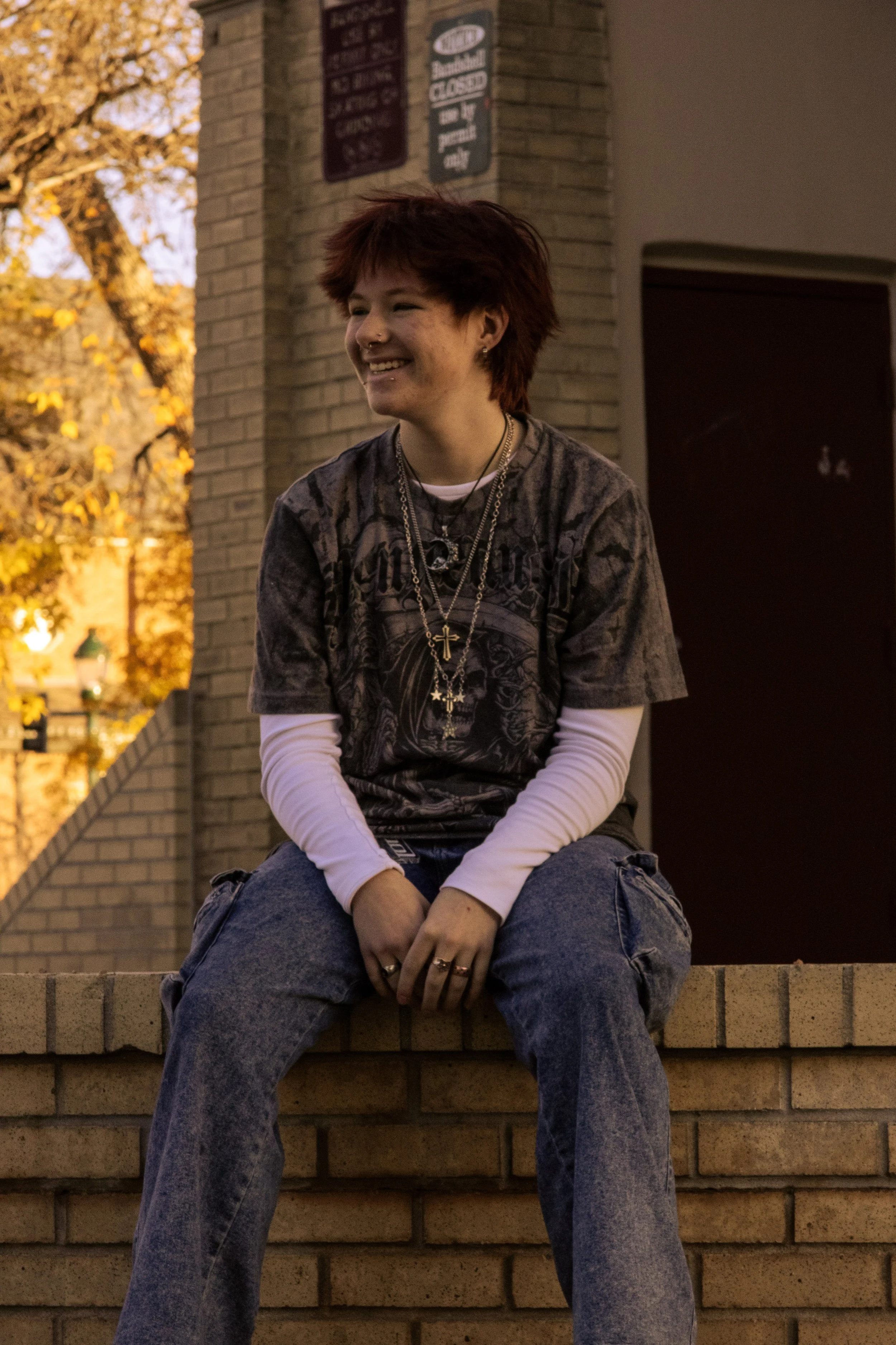 Young woman with short red hair sitting on a brick ledge at dusk, smiling and wearing layered clothing with jewelry, with a tree and building behind her.