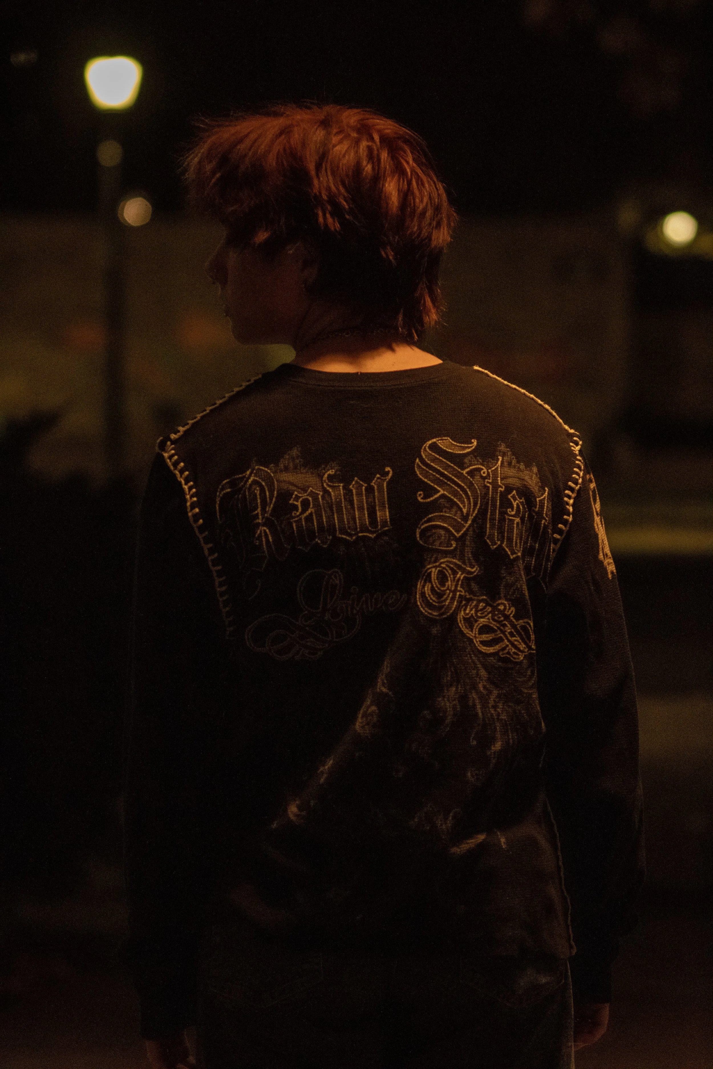 A person with short, layered hair, wearing a black shirt with ornate gold lettering and designs, standing in a dark setting with a few blurred streetlights or light sources in the background.