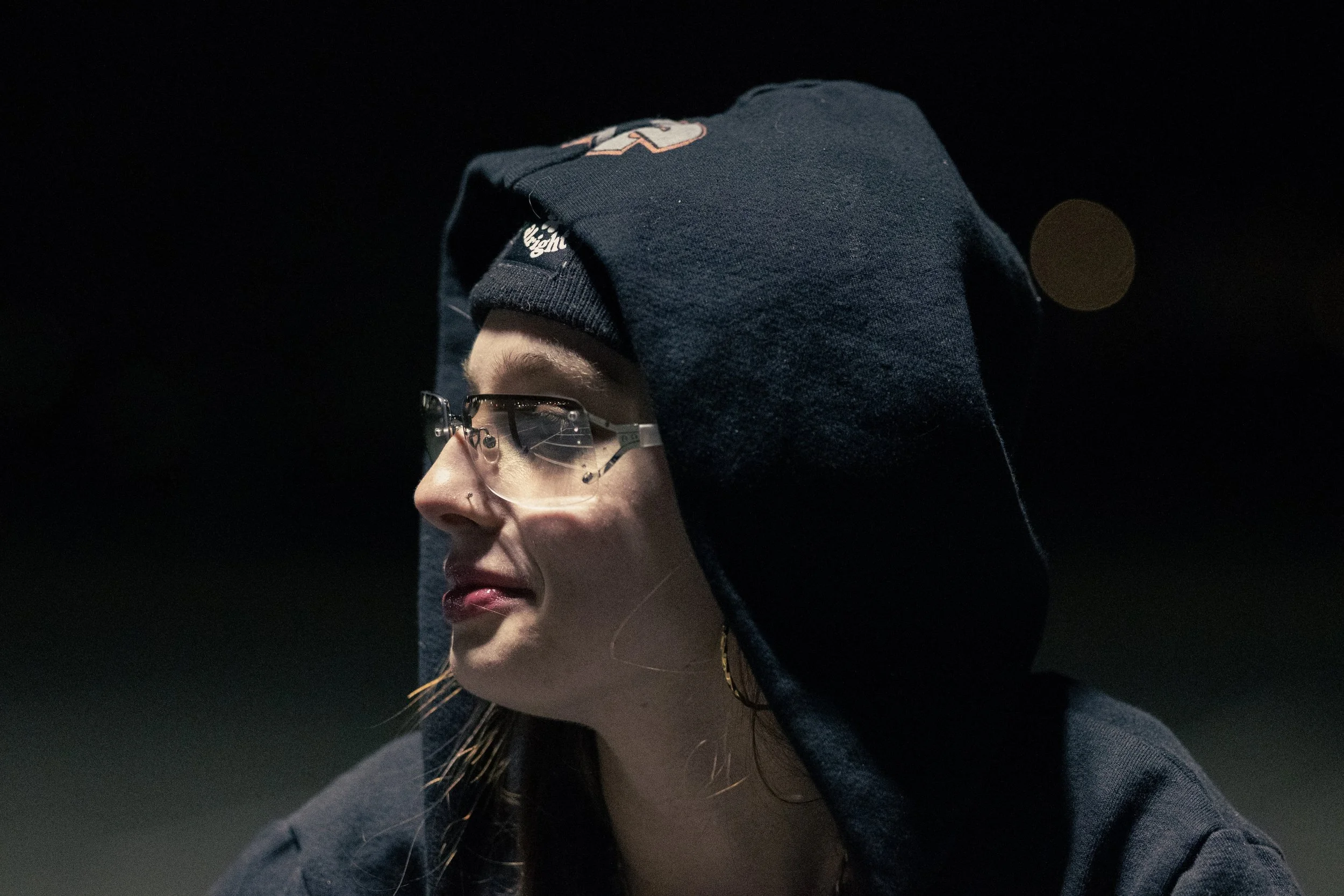 A person wearing glasses and a navy blue hoodie with the hood over their head, lit in a dark environment.