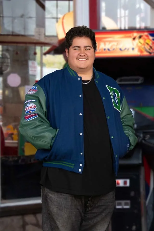 A young man with dark hair, smiling, wearing a blue and green varsity jacket with patches, standing inside a colorful arcade or entertainment center.