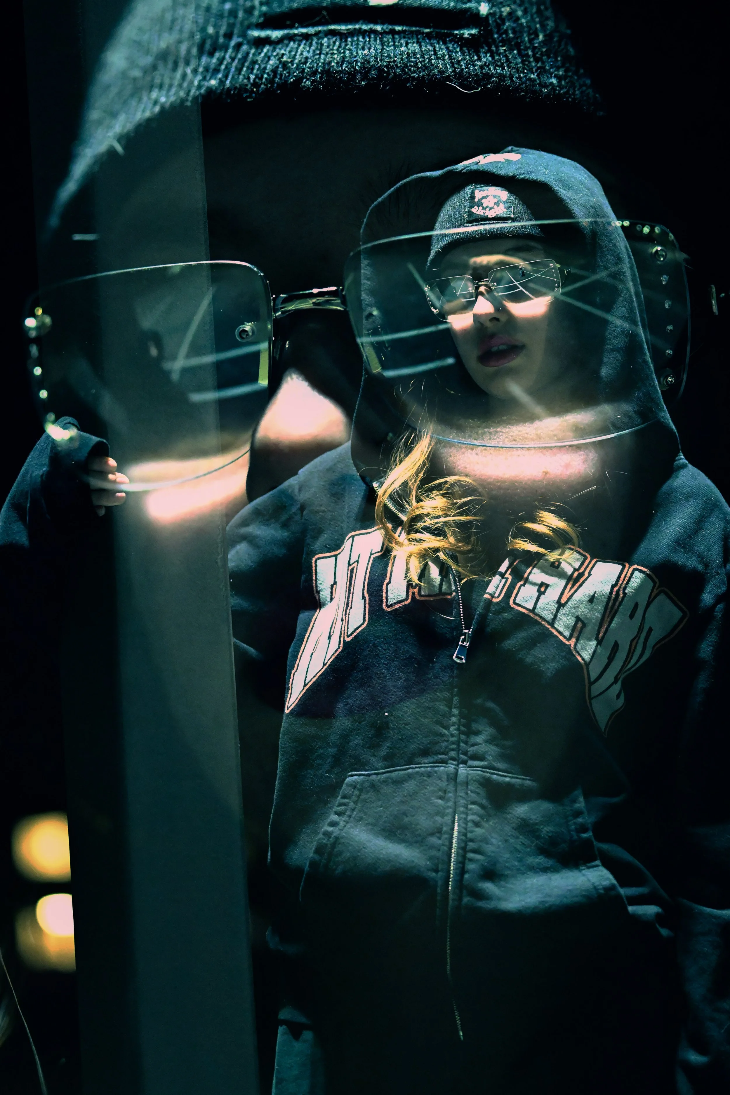 A woman wearing a hoodie, sunglasses, and a beanie hat, looking into a reflective surface with neon lighting and dark background.