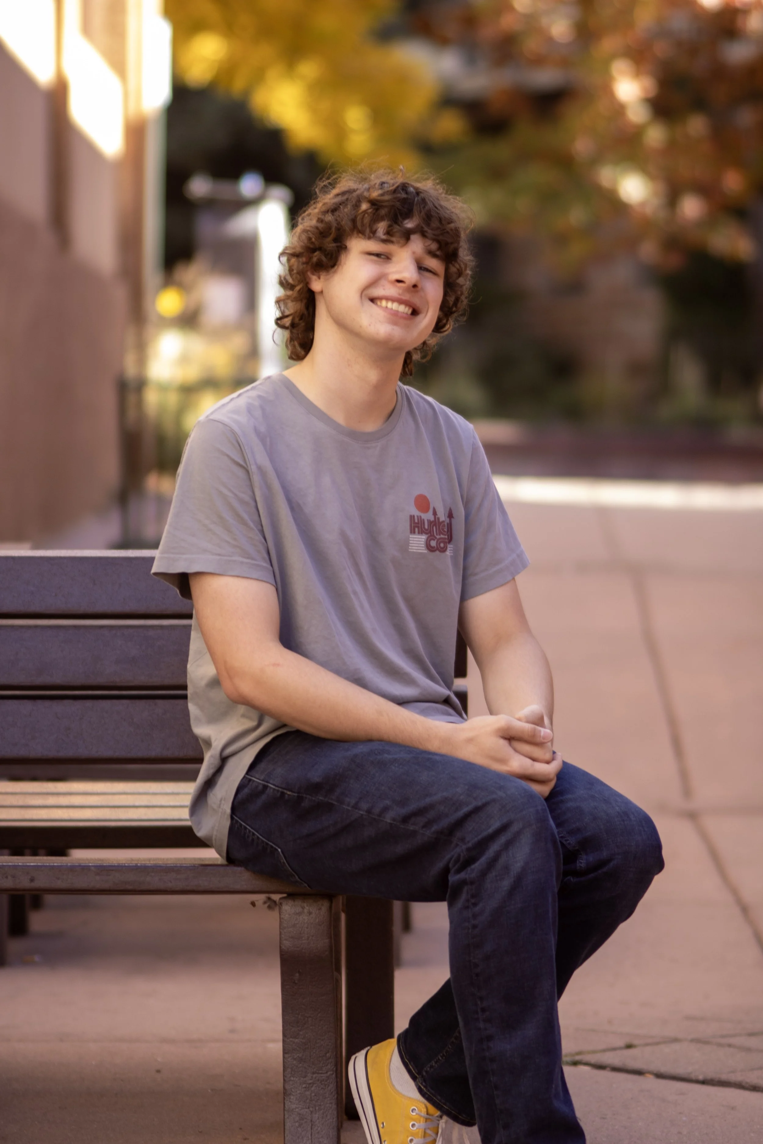 A young man with curly brown hair sitting on a park bench, smiling at the camera, wearing a gray t-shirt with a logo, dark jeans, and yellow sneakers, with autumn trees in the background.