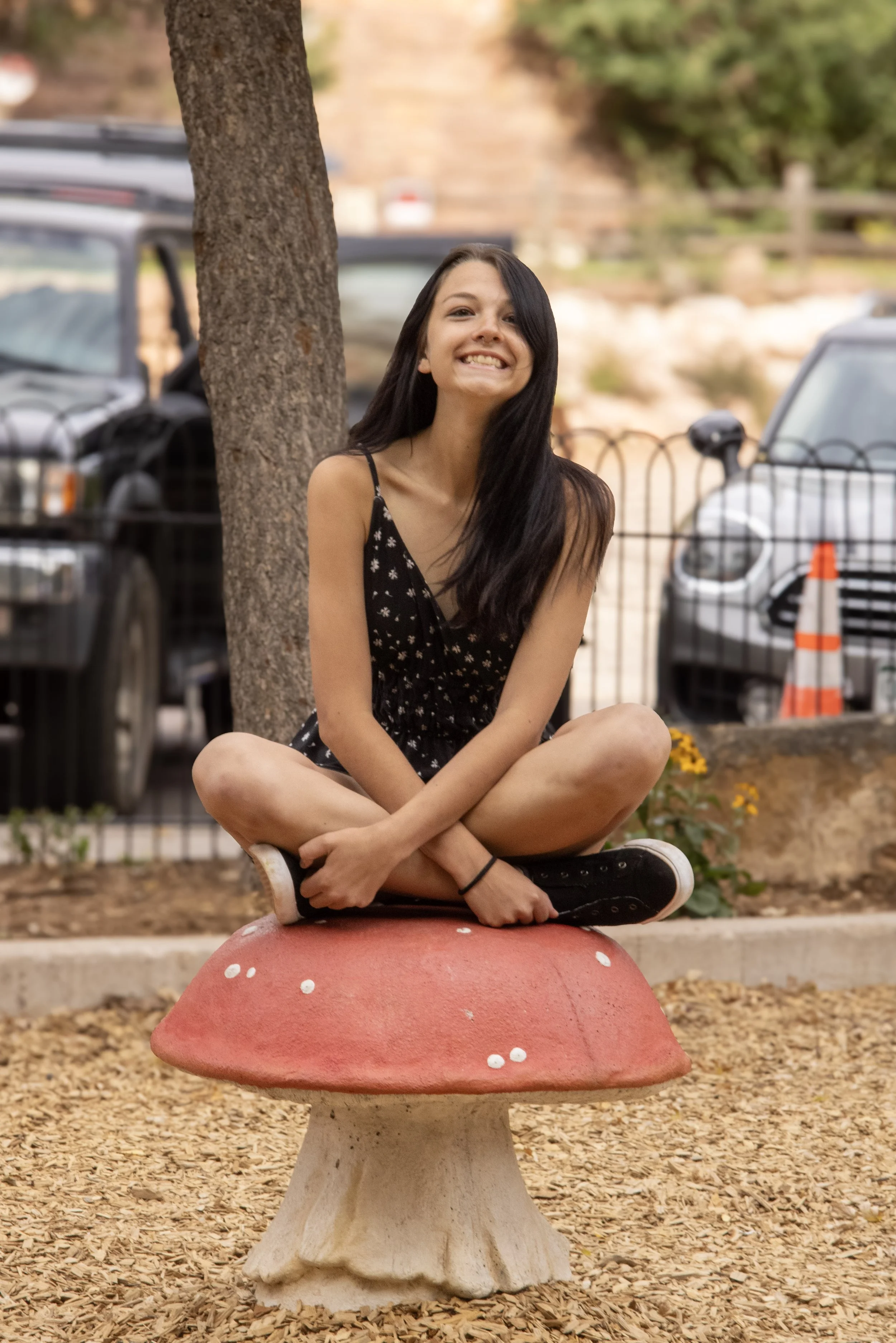 A young woman with long dark hair sitting cross-legged on a mushroom-shaped sculpture in a park, smiling at the camera. She is wearing a black dress with white floral patterns and black sneakers. The background shows parked cars and a blurred park setting with trees and a fence.
