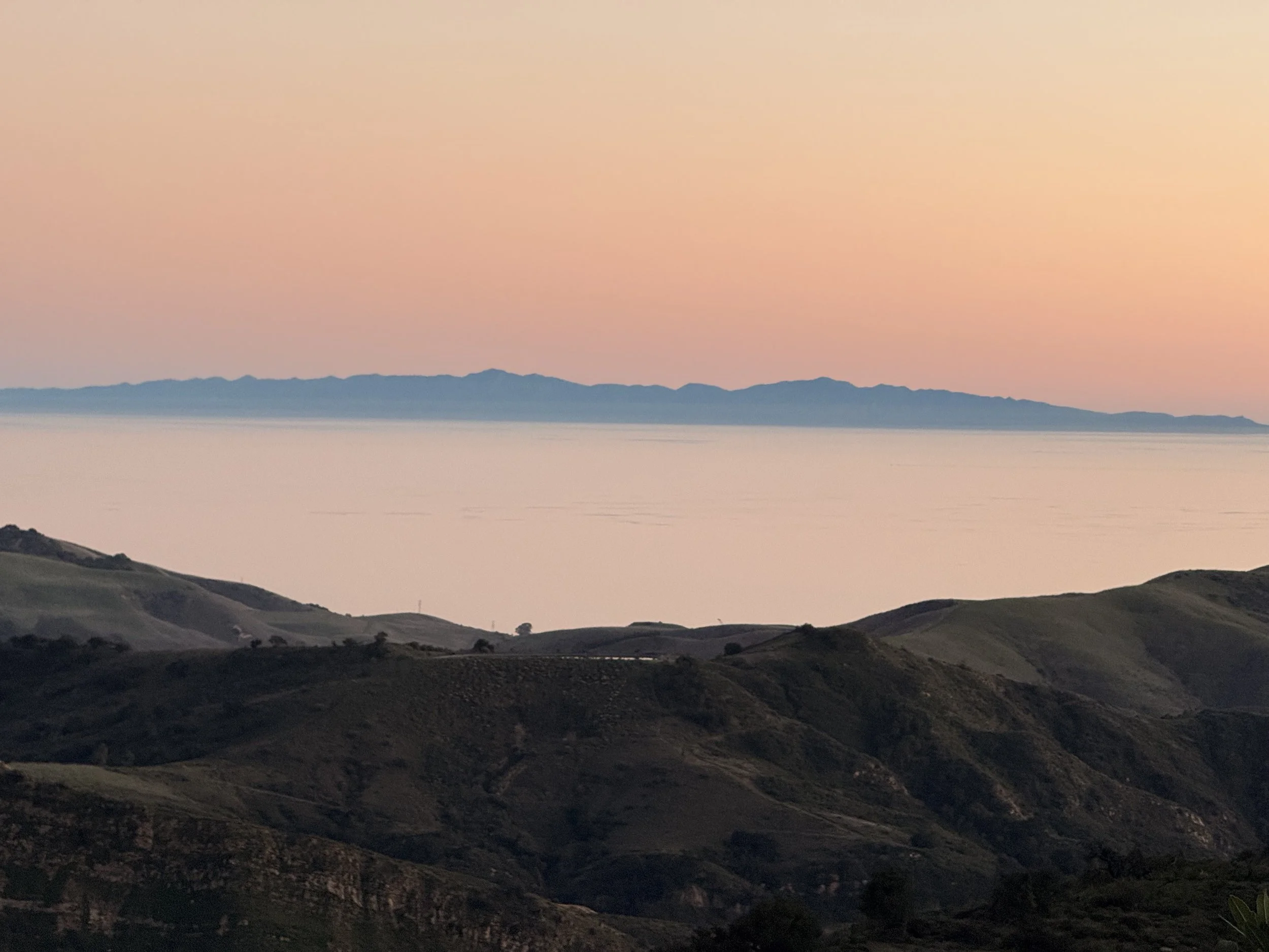 A landscape scene at sunset showing hills and mountains in the distance, with the sky displaying a gradient of soft pastel colors from light pink to peach.