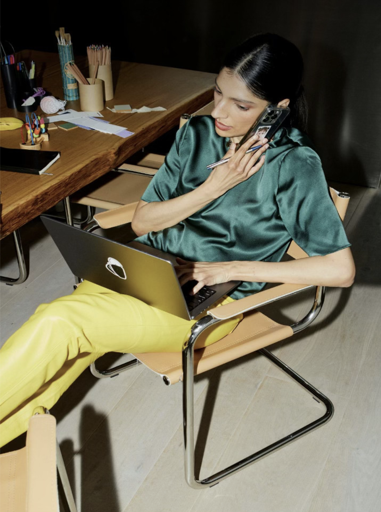 Woman sitting in a chair working on a laptop and talking on the phone in an office or workspace setting with various office supplies on the desk nearby.