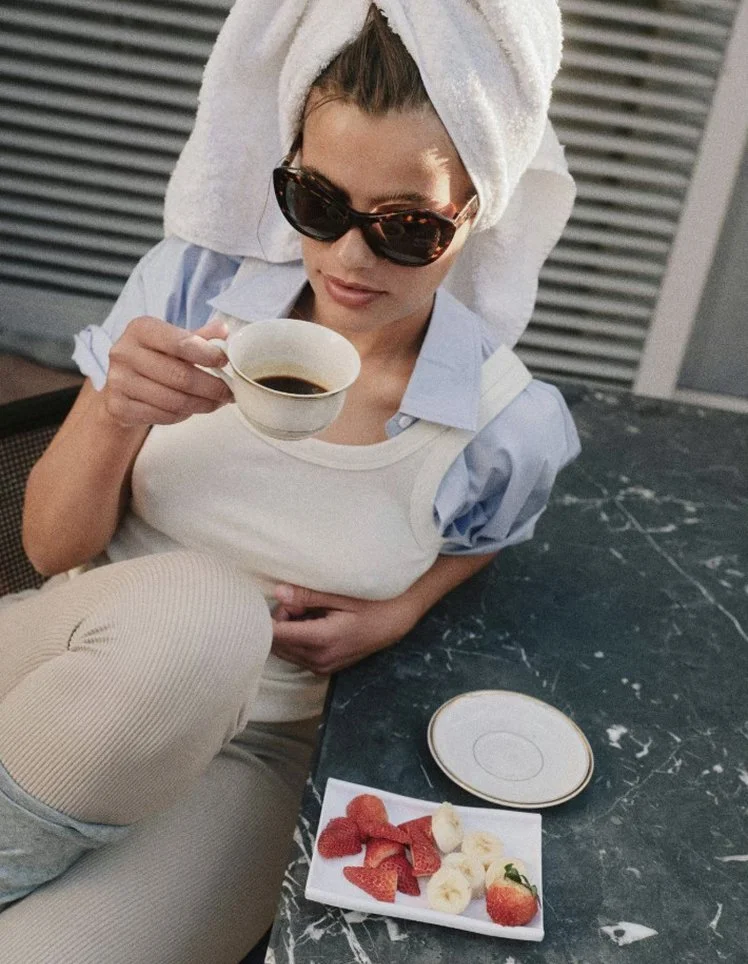 A woman with sunglasses and a towel wrapped around her head, sitting at a marble table, holding a cup of coffee, with a plate of strawberries and bananas nearby.