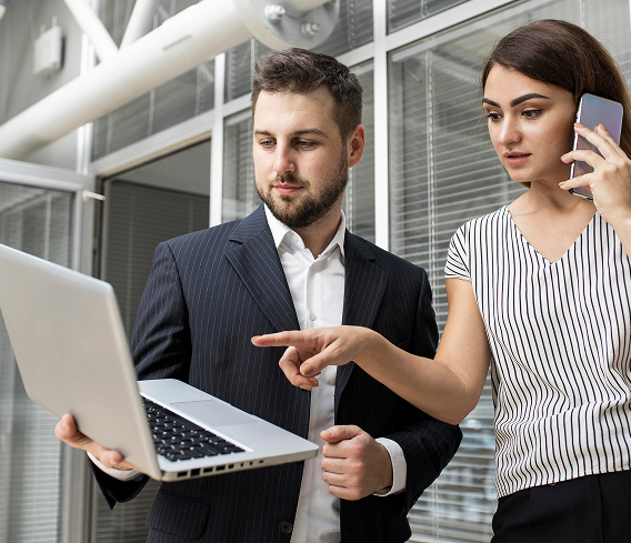 Two professionals, a man in a business suit and a woman in a striped top, are looking at a laptop outside a modern building. The woman is talking on her phone and pointing at the laptop.