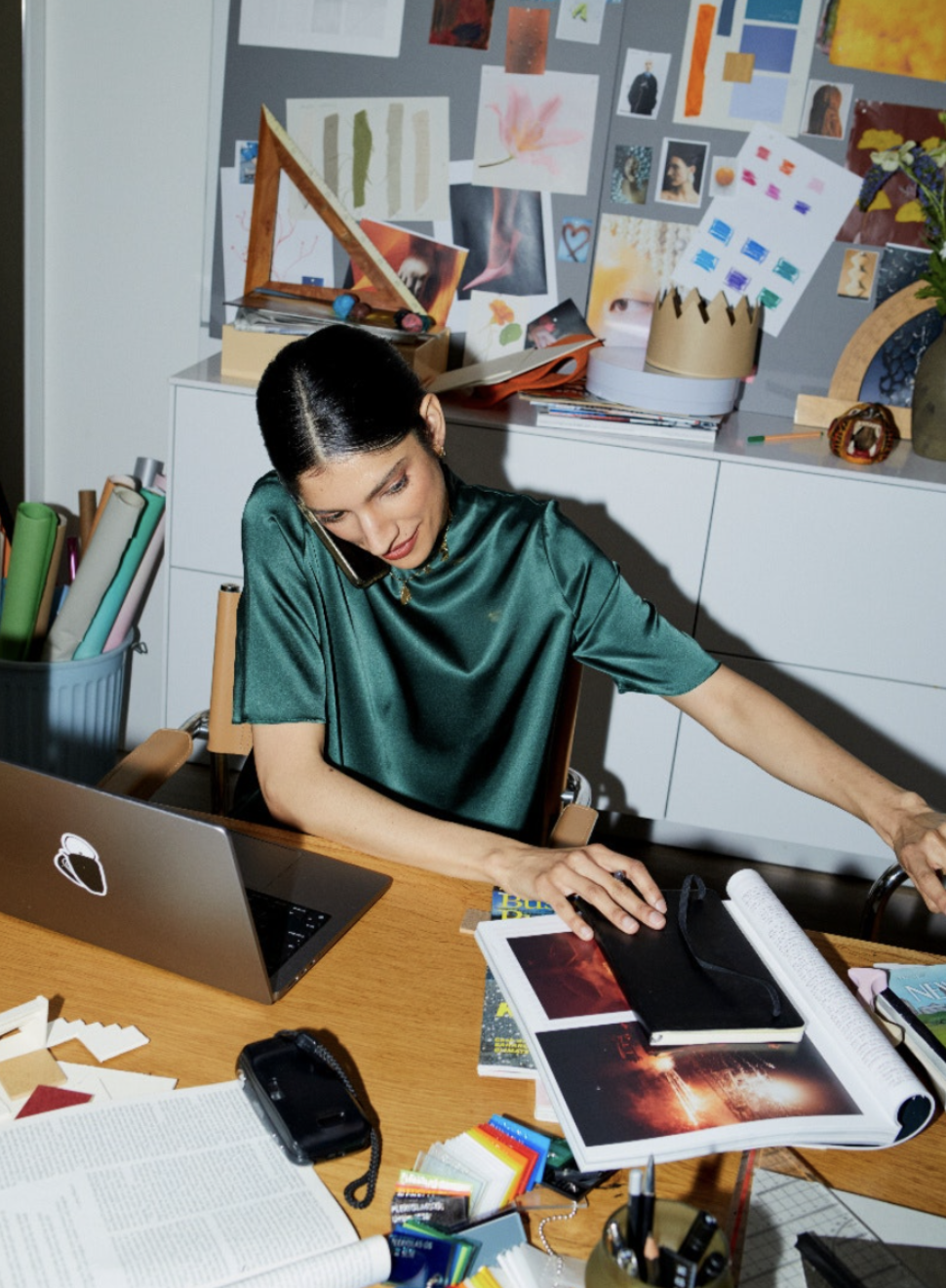 A woman sitting at a cluttered desk talking on the phone, with various art supplies, magazines, and a laptop, in a room with a bulletin board filled with artwork.