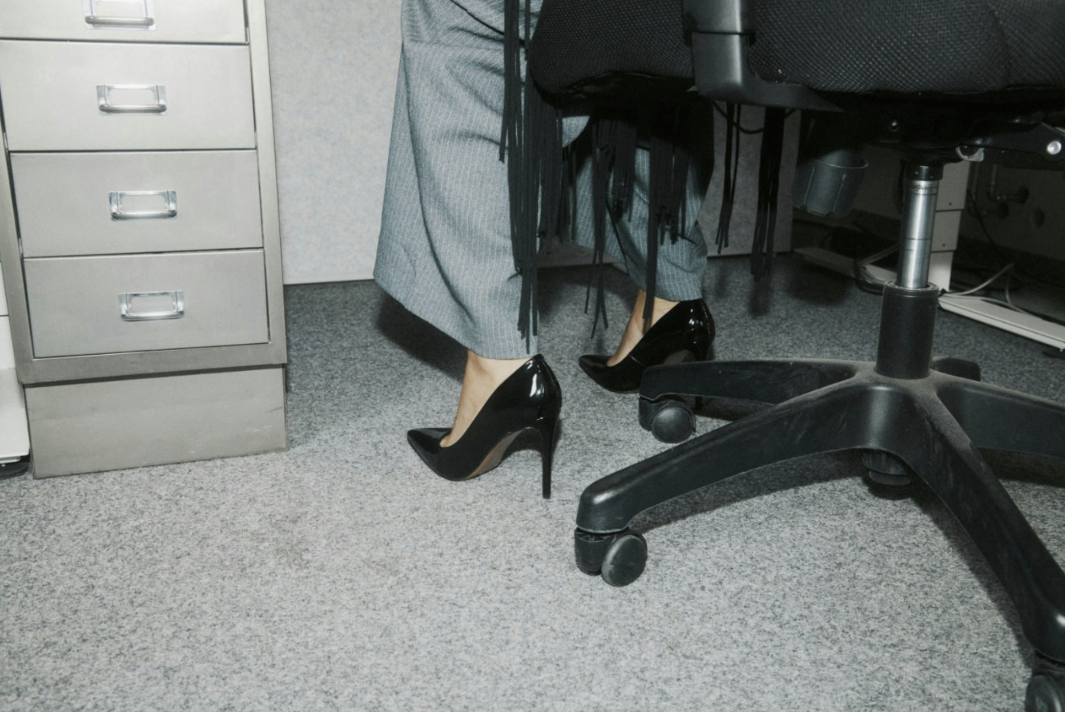 A person sitting at an office desk wearing high-heeled shoes and pinstripe pants, with part of the office environment, including filing cabinets, a chair, and equipment visible.