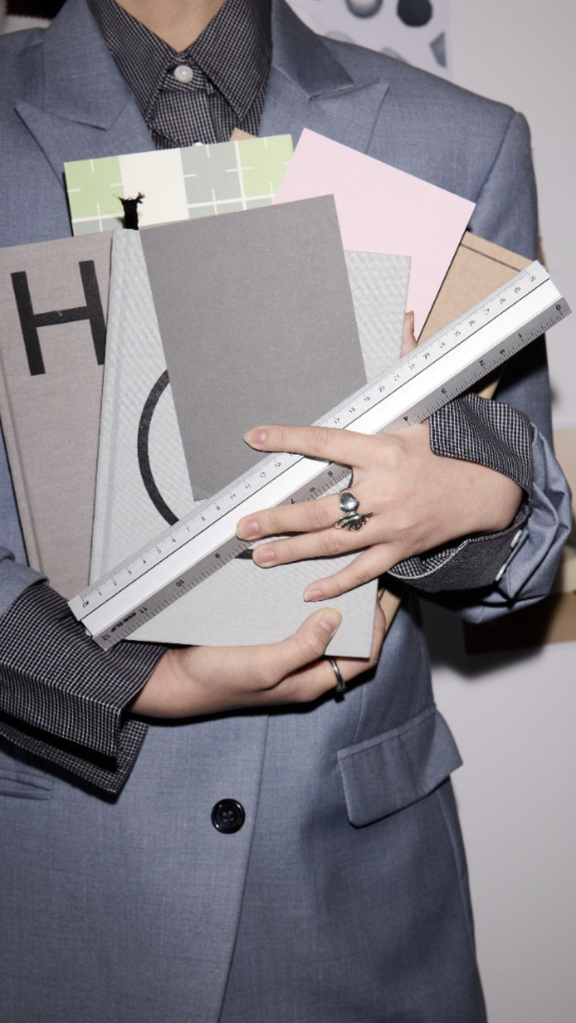 Person holding several colored books or notebooks and a white ruler, dressed in a grey suit and patterned shirt.