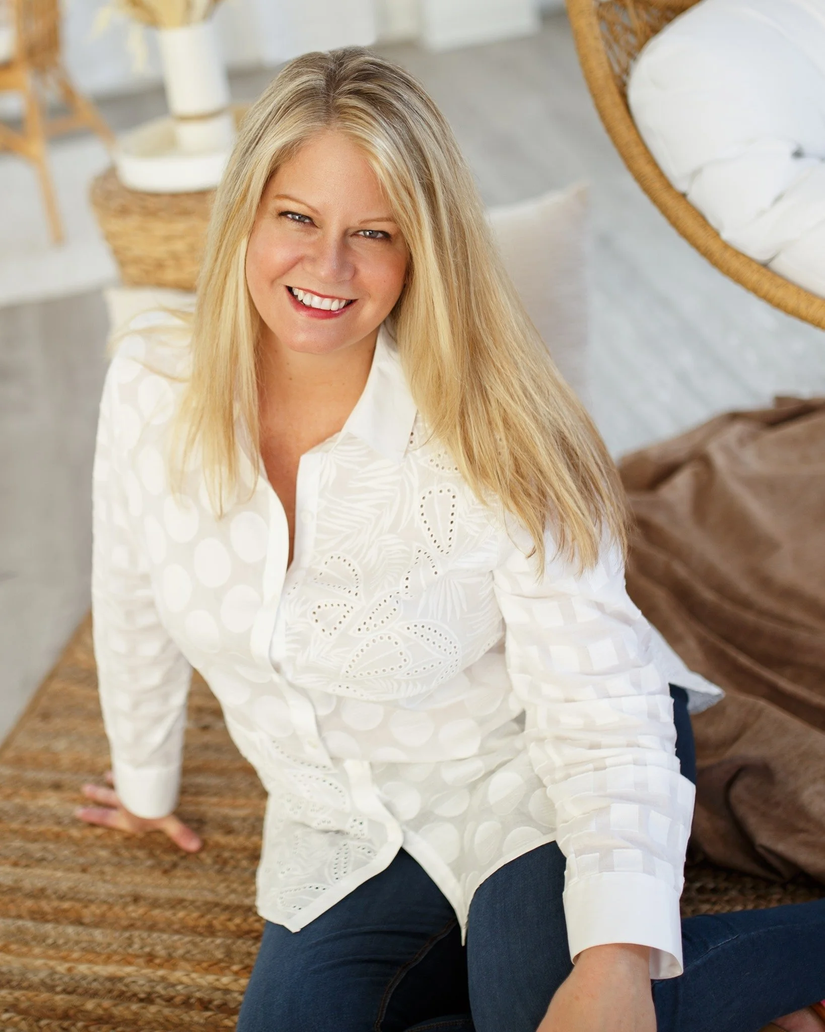 Woman smiling in white blouse and dark jeans sitting on a carpeted floor with a rattan chair and shelf in the background.